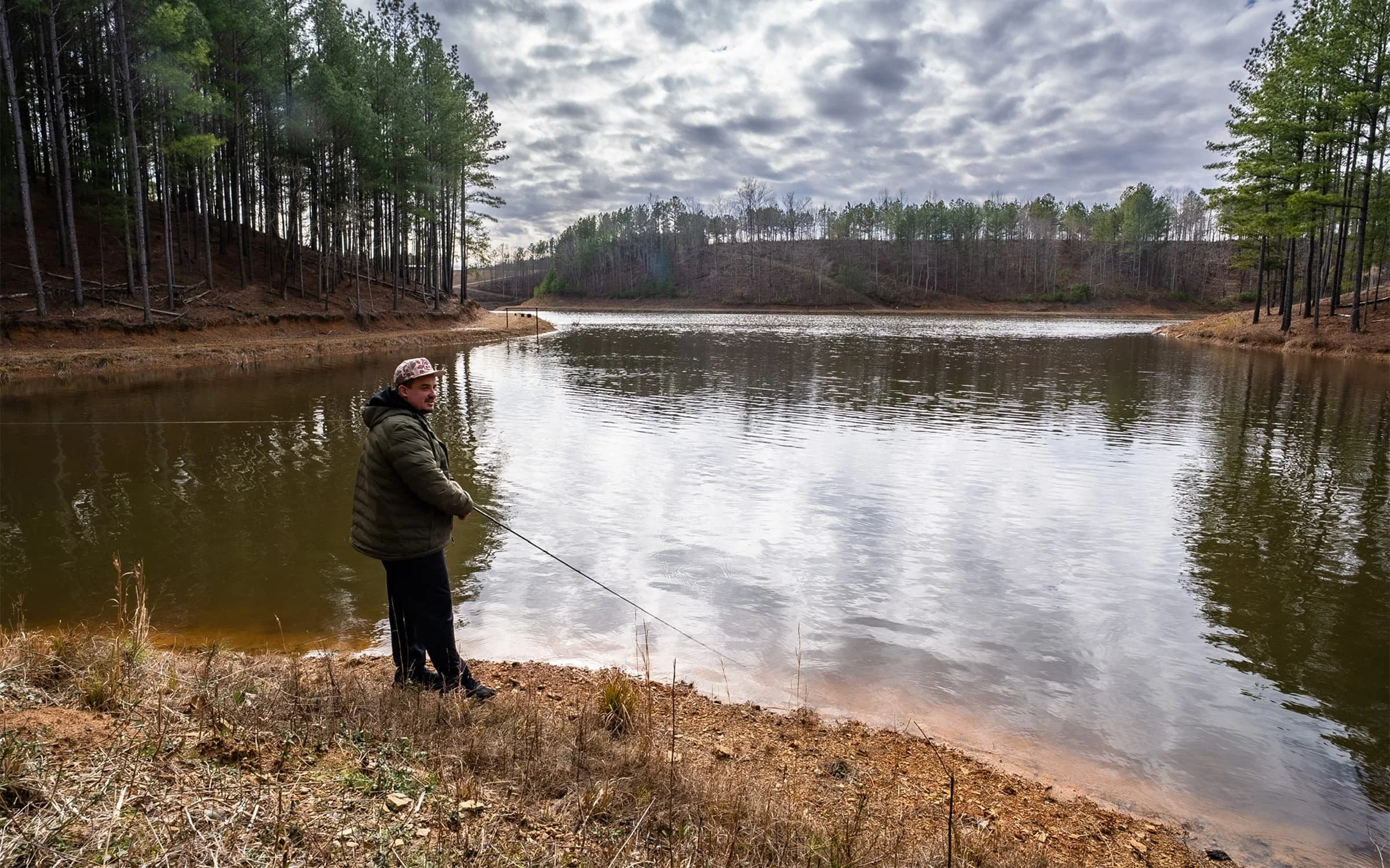 A man stands by a lake, fishing with a rod. The lake is surrounded by trees, with a cloudy sky above.