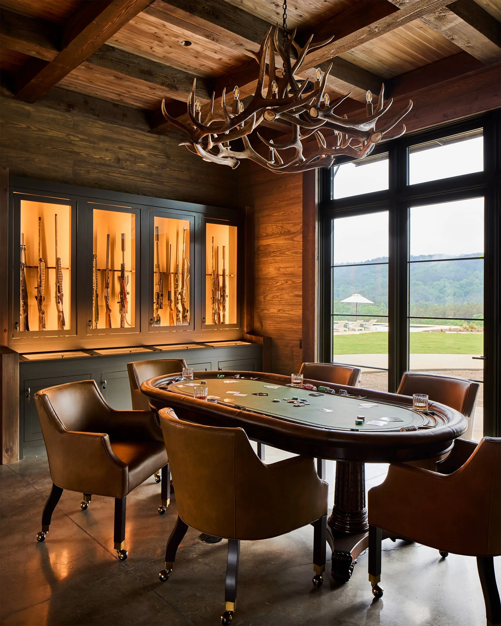 A poker game setup with a round table, four leather chairs, and playing chips and cards on the table. The room has wooden walls and ceiling, with a large window revealing an outdoor landscape with lounge chairs and a patio umbrella.