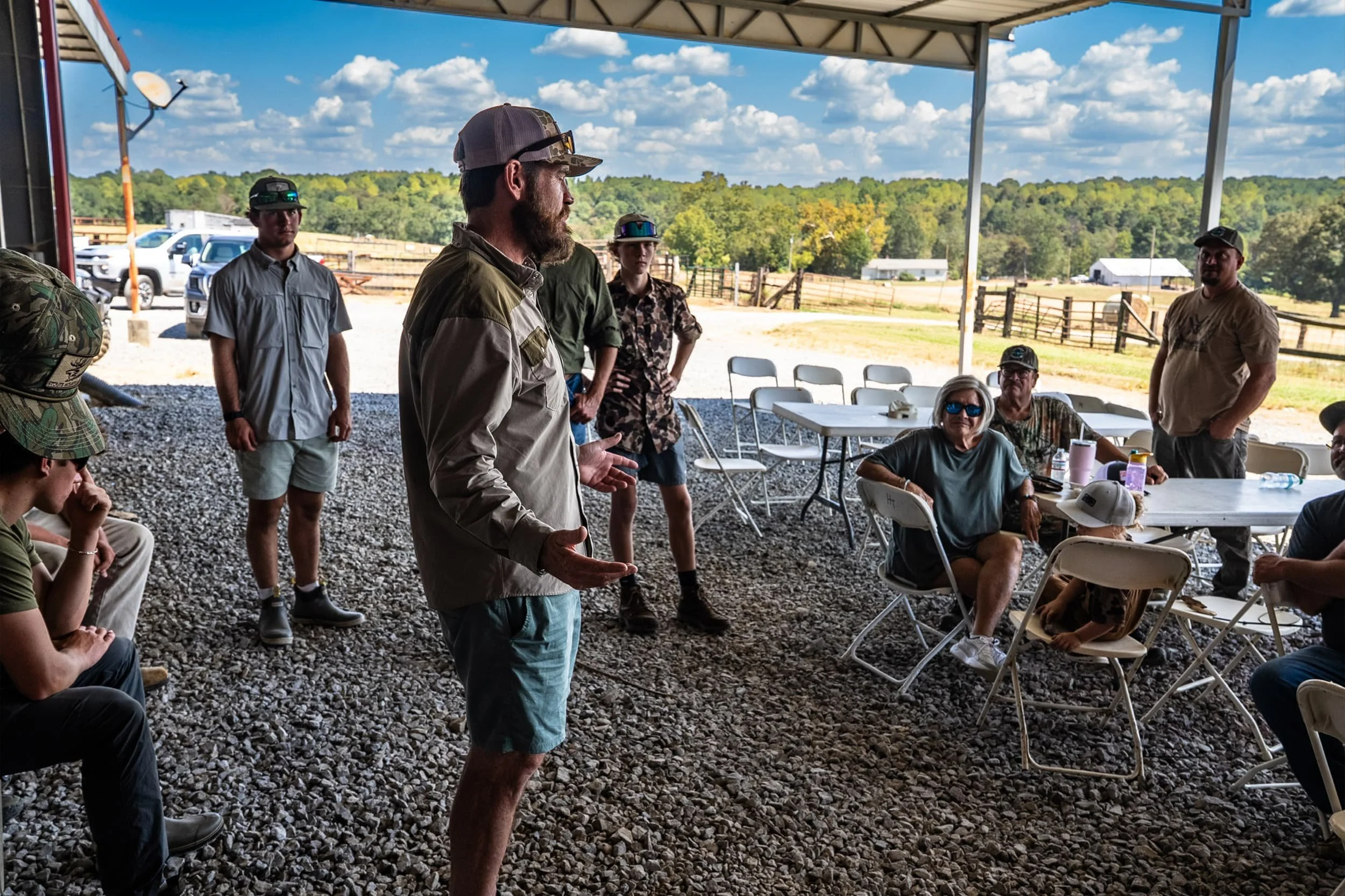 A man with a beard, wearing a baseball cap and casual clothing, is speaking to a group of people under a large open-sided shelter. Several audience members are seated in white folding chairs, and some are wearing hats and sunglasses. The background shows a rural landscape with green trees, a few buildings, and a partly cloudy sky.