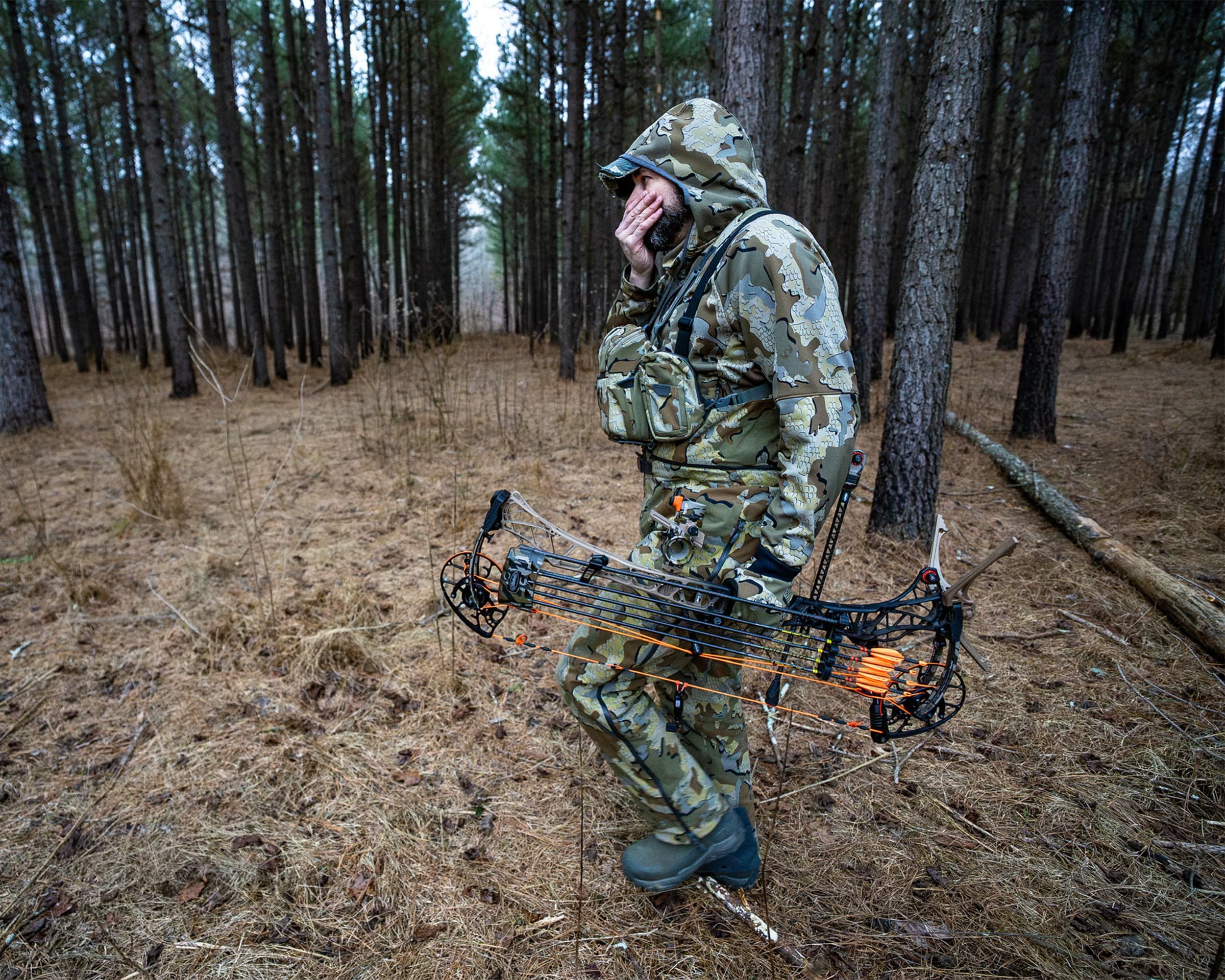 A man dressed in camouflage outdoor gear walking through a forest, carrying a compound bow.