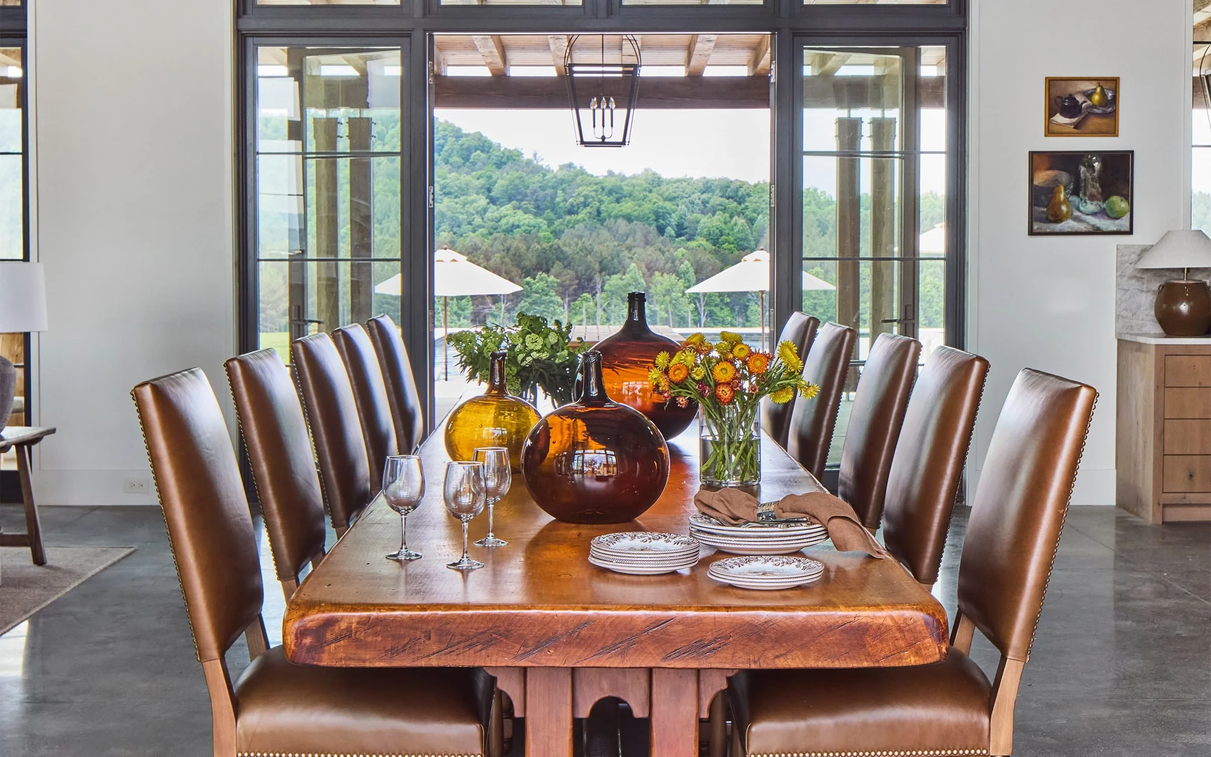 Interior of a dining room with a wooden table set with glassware, napkins, and a floral centerpiece. The background features large glass doors open to a patio with outdoor umbrellas and lush green hills.