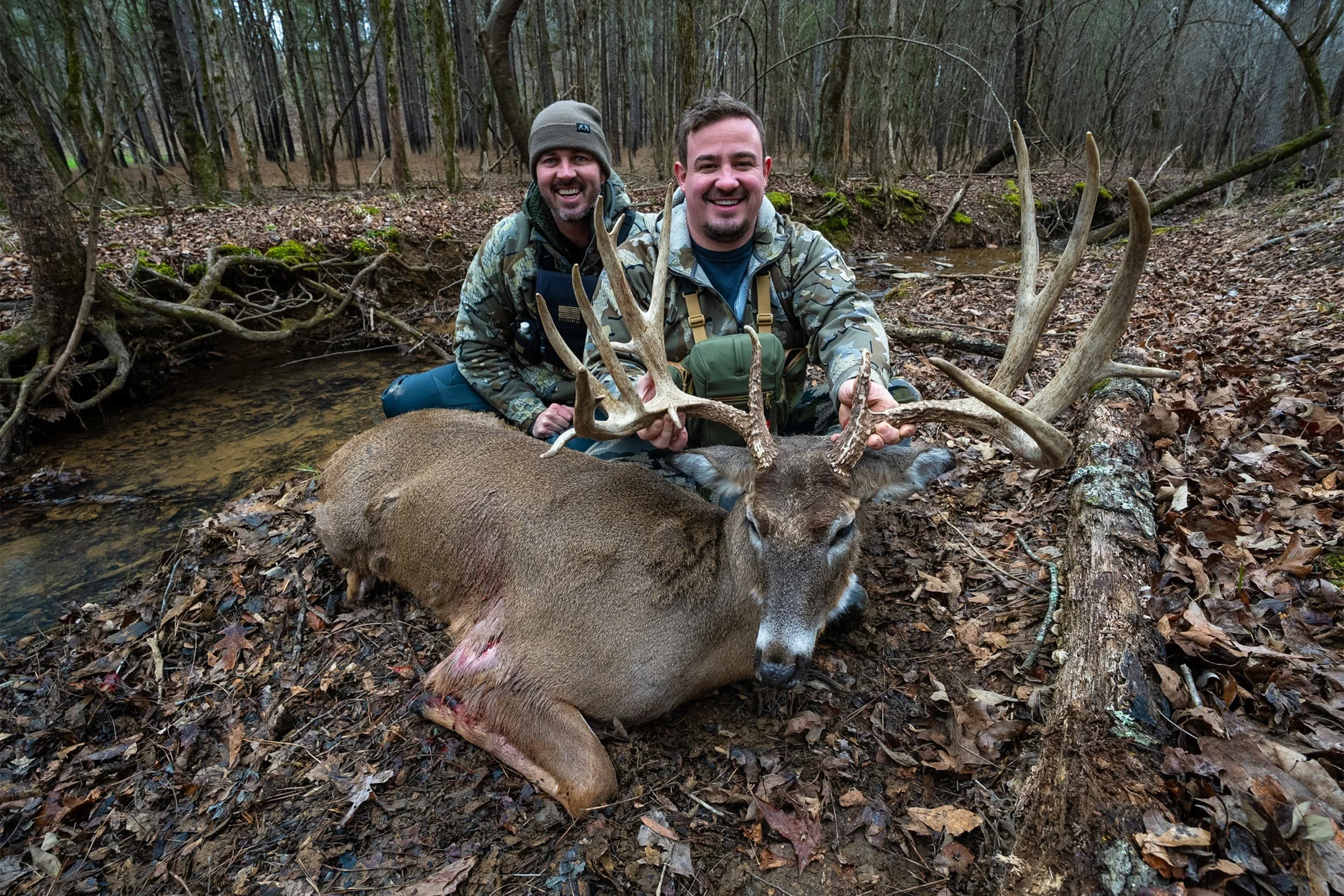 Two smiling men in camouflage jackets kneel in a forested area by a small creek, holding the antlers of a large deer that is lying on the ground with its eyes closed.