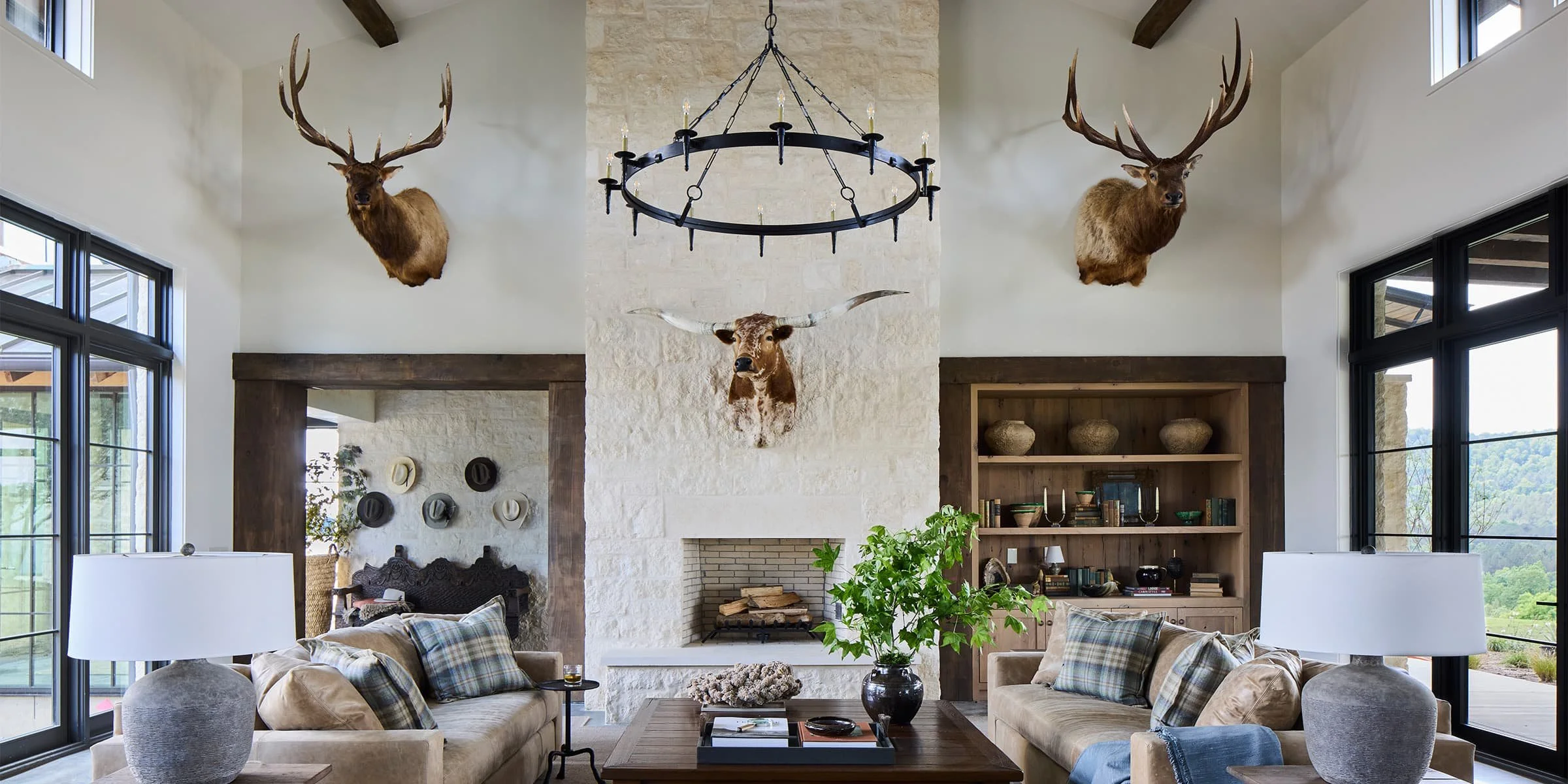 Living room with a white fireplace, ceiling beams, and large windows, decorated with mounted animal heads including a central longhorn skull and two deer with antlers above the fireplace.