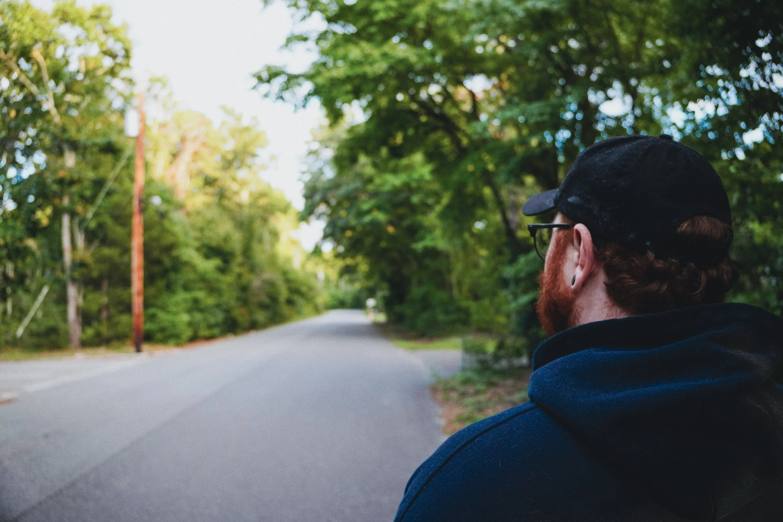 A man with a beard and glasses is seen from behind, wearing a black cap and hoodie, standing on a tree-lined street during daytime.