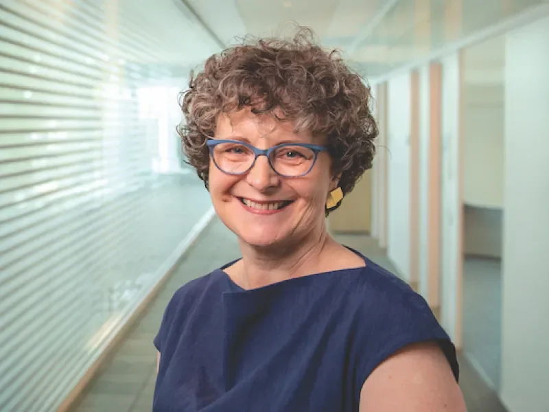 A smiling older woman with curly hair, blue glasses, and gold earrings standing in a modern hallway.