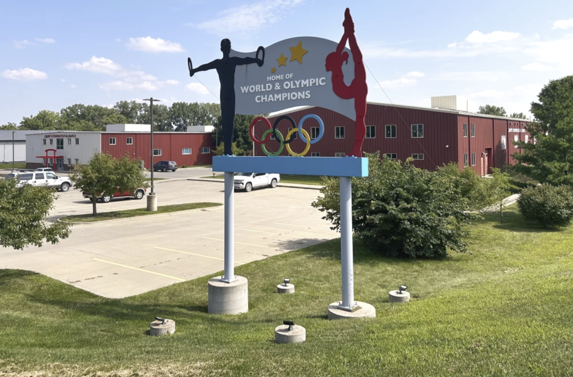 Sign with figures of a gymnast and a diver, Olympic rings, and text reading "Home of World & Olympic Champions", in a parking lot with trees and a red and white building in the background.