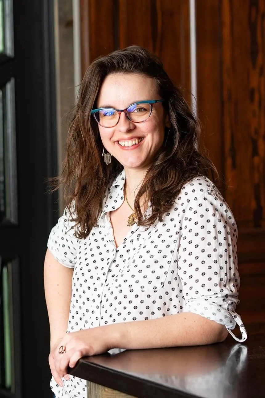 Smiling woman with wavy brown hair, wearing blue and pink glasses, a polka dot shirt, and earrings, standing indoors near a wooden wall and a bookshelf.