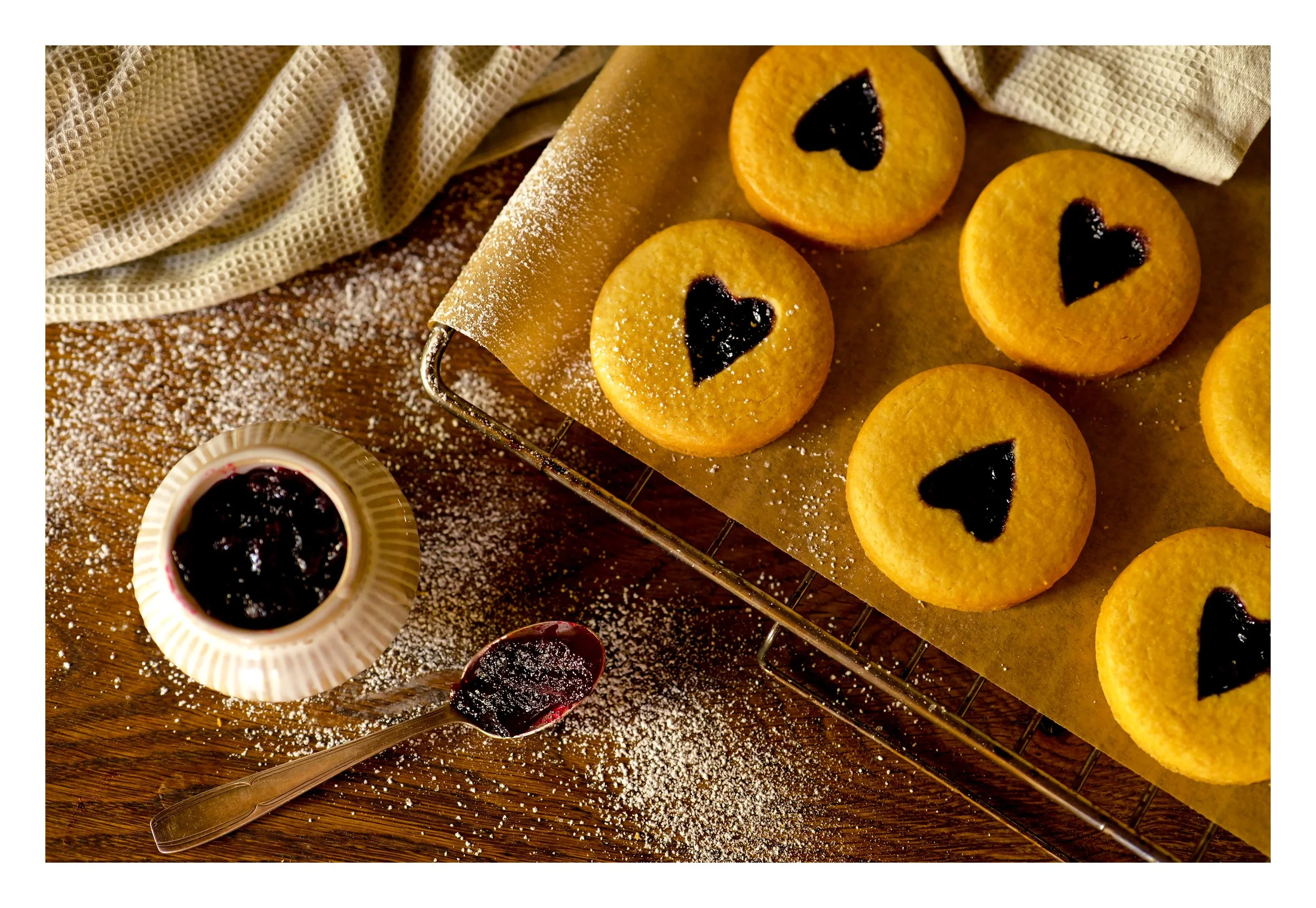 Biscuits ronds avec un cœur en confiture, saupoudrés de sucre glace, avec un bol de confiture de fruits rouges à côté, sur une surface en bois et un tissu beige en arrière-plan.