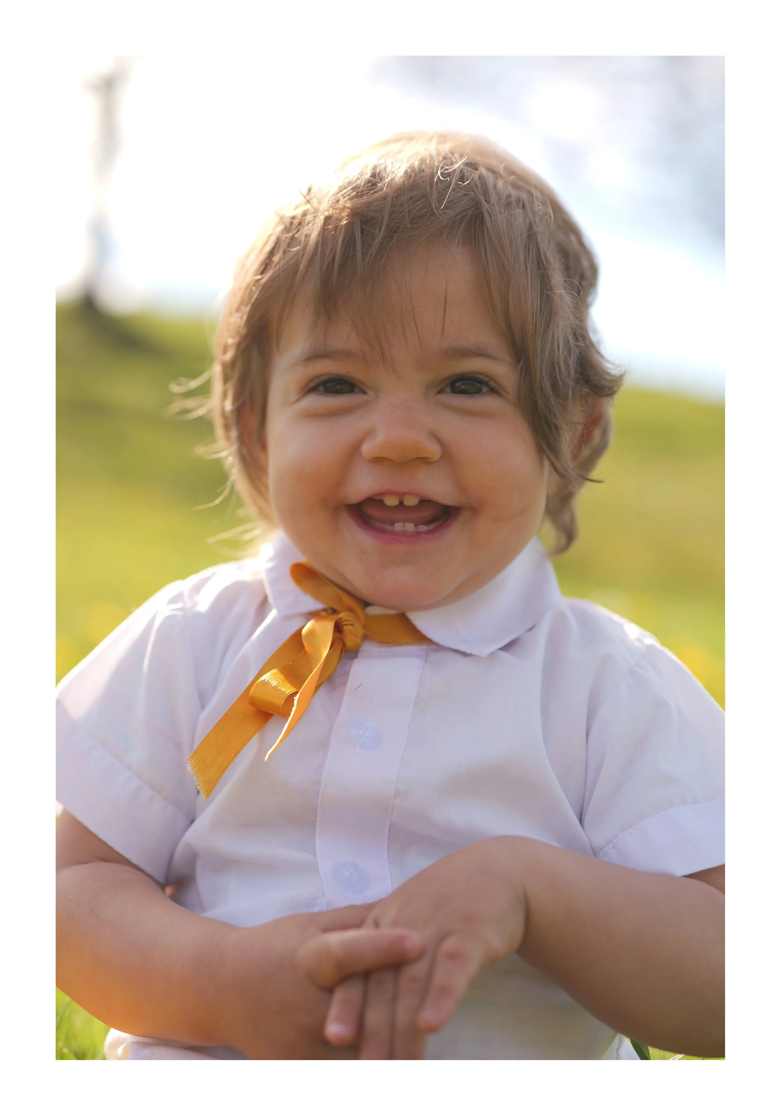 Jeune enfant souriant dans un champ en plein air, portant une chemise blanche avec un nœud papillon orange et montant ses mains ensemble.