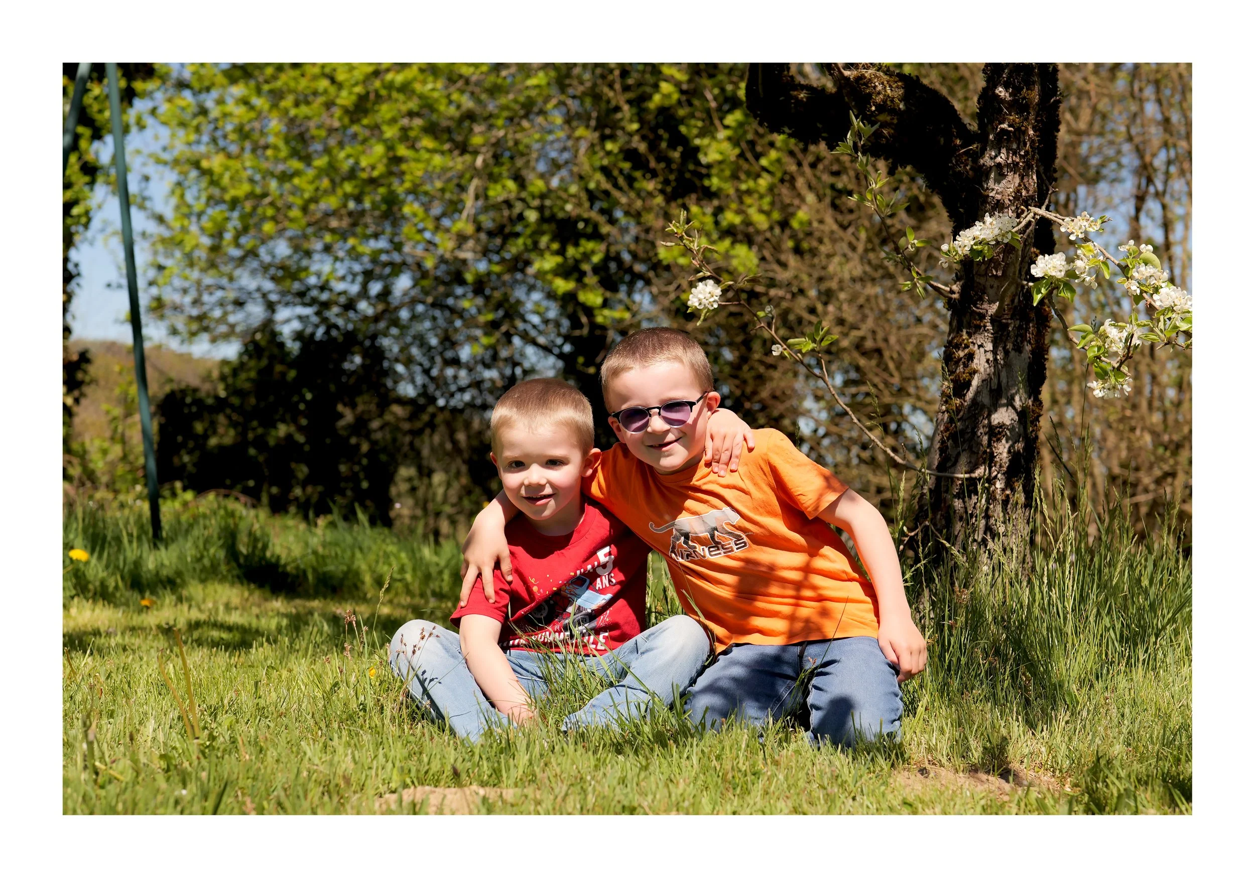 Deux enfants souriants assis sur l'herbe dans un parc, entourés d'arbres en fleurs, ensoleillement printanier.