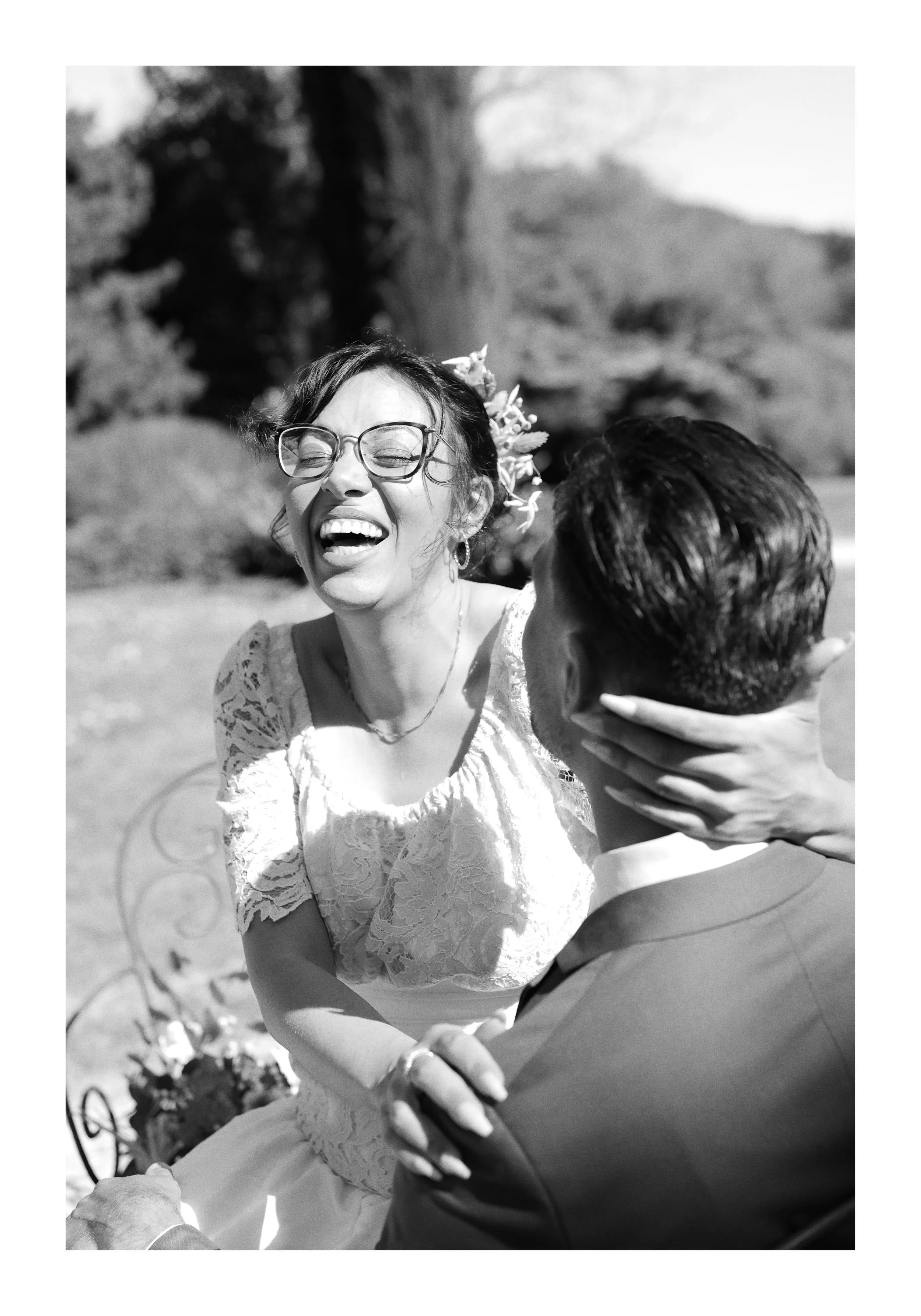 Une femme souriante portant des lunettes, vêtue d'une robe en dentelle, semble danser ou se déplacer joyeusement lors d'une célébration extérieure, avec un homme dont le dos est visible, en costume, l'accompagnant.