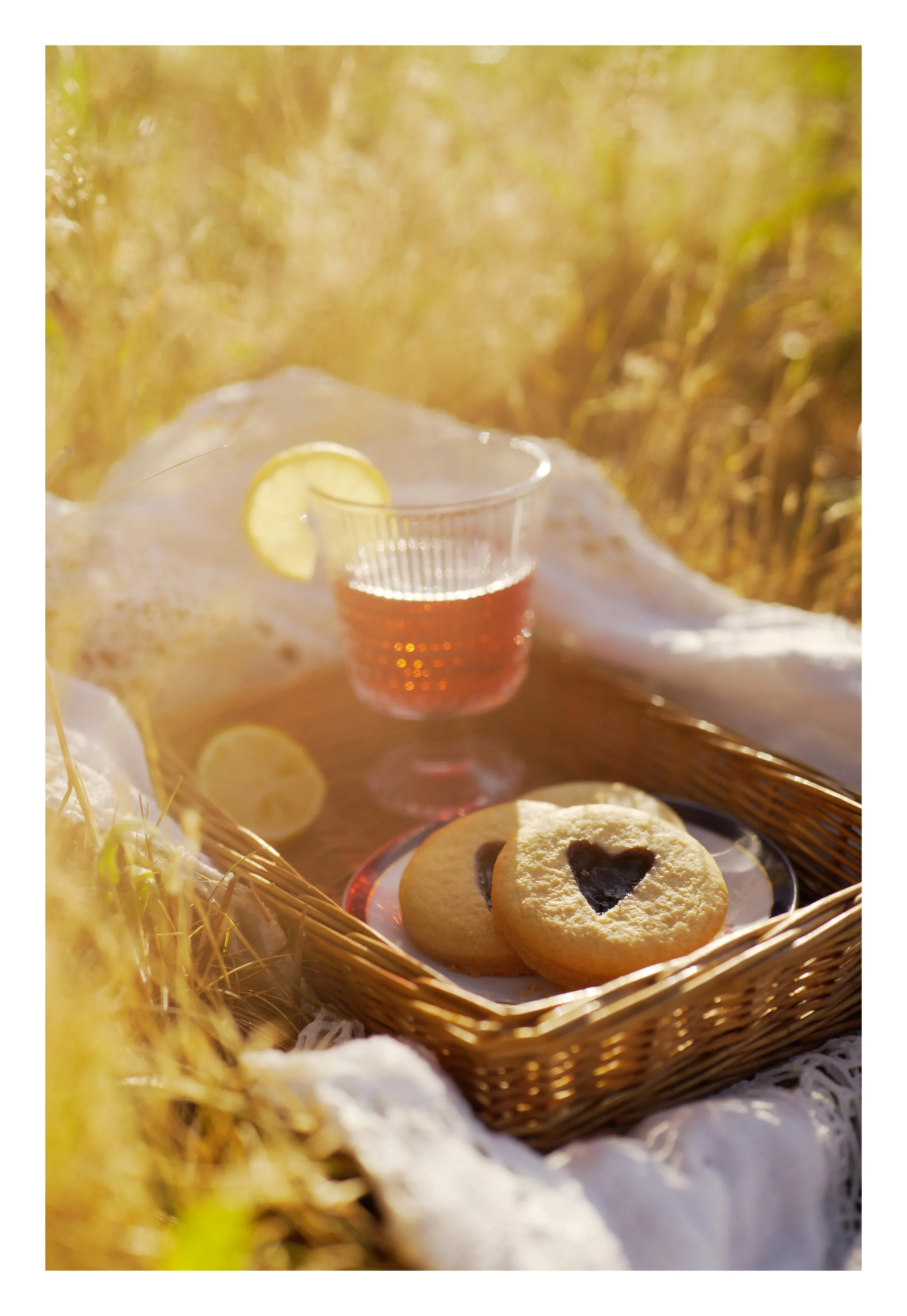 Une tasse de thé ou de boisson chaude avec une tranche de citron, entourée de biscuits avec un cœur au chocolat, dans une corbeille en osier, en plein air, avec un fond flou de champs dorés au soleil.