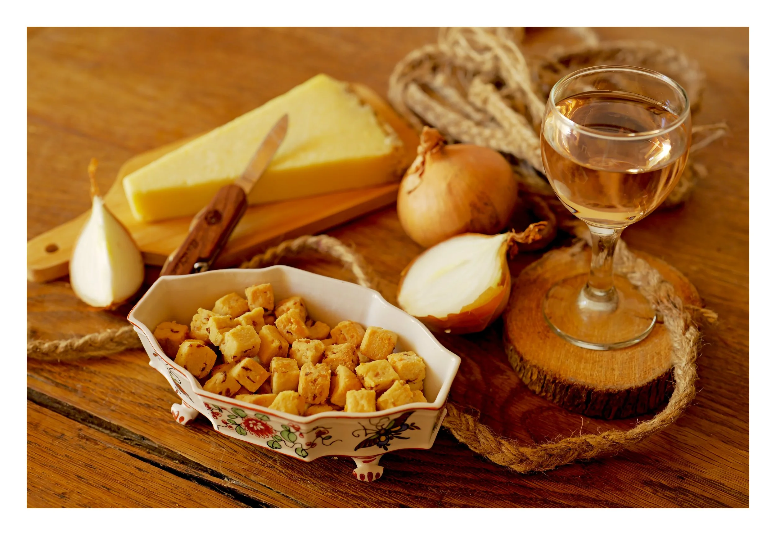 Une assiette remplie de croûtons, un morceau de fromage, un oignon, une bouteille de vin blanc, et des ingrédients de cuisine sur une planche en bois.