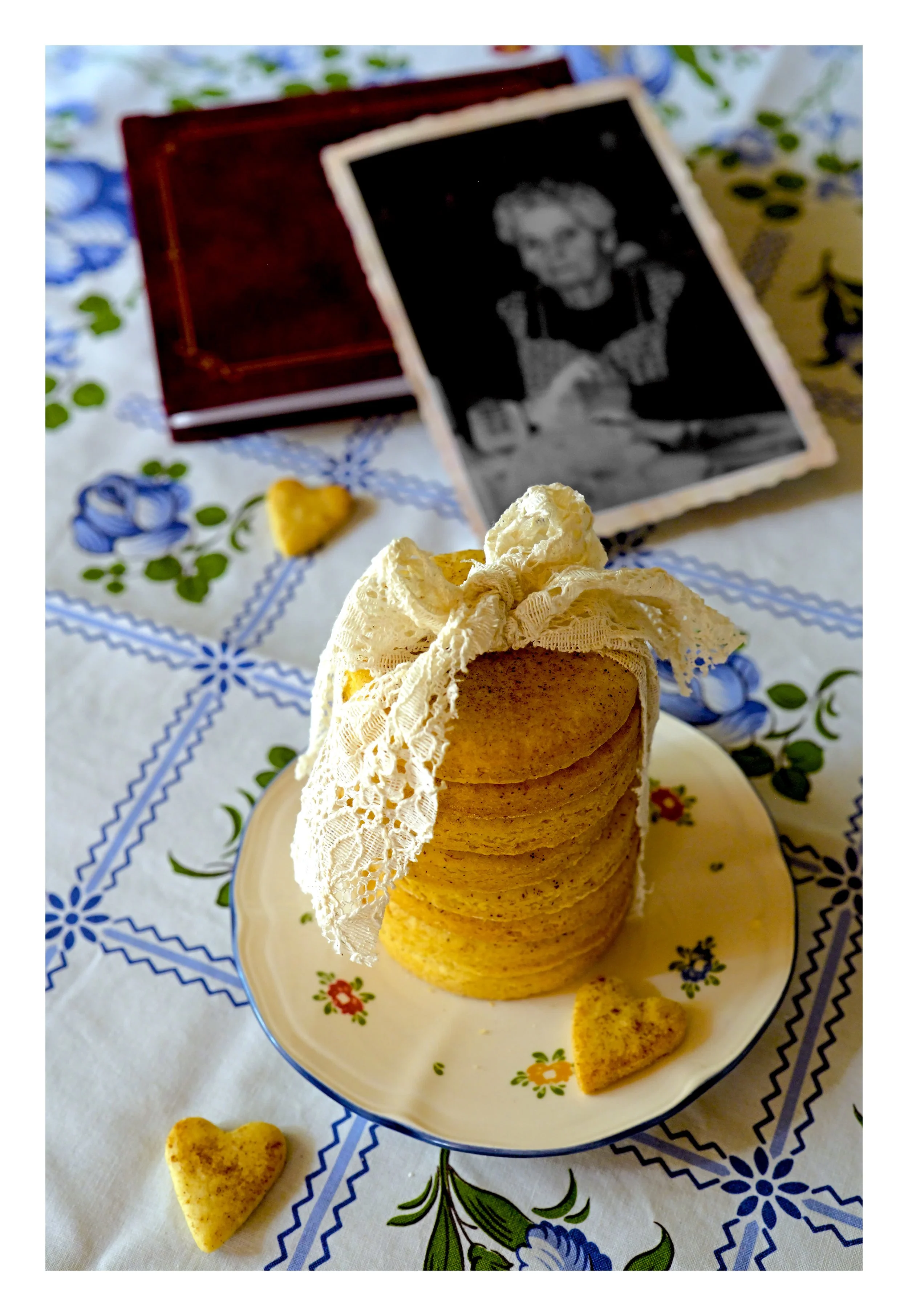 Une assiette avec une petite pile de cookies de forme heart avec une décoration en dentelle. Au-dessus, un décor de table avec un motif floral. En arrière-plan, une photo en noir et blanc d'une femme dans un cadre en bois et un petit cœur en biscuit