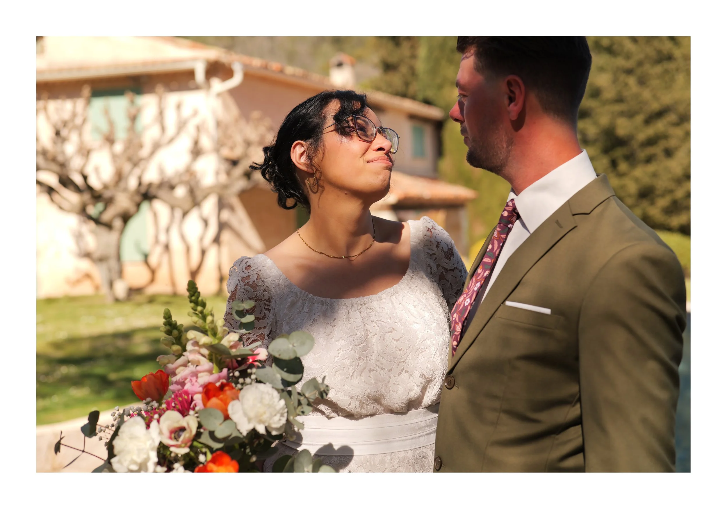 Un couple lors de leur mariage, la femme porte une robe en dentelle blanche et donne un bouquet de fleurs colorées. Ils se regardent tendrement dans un jardin en plein air.