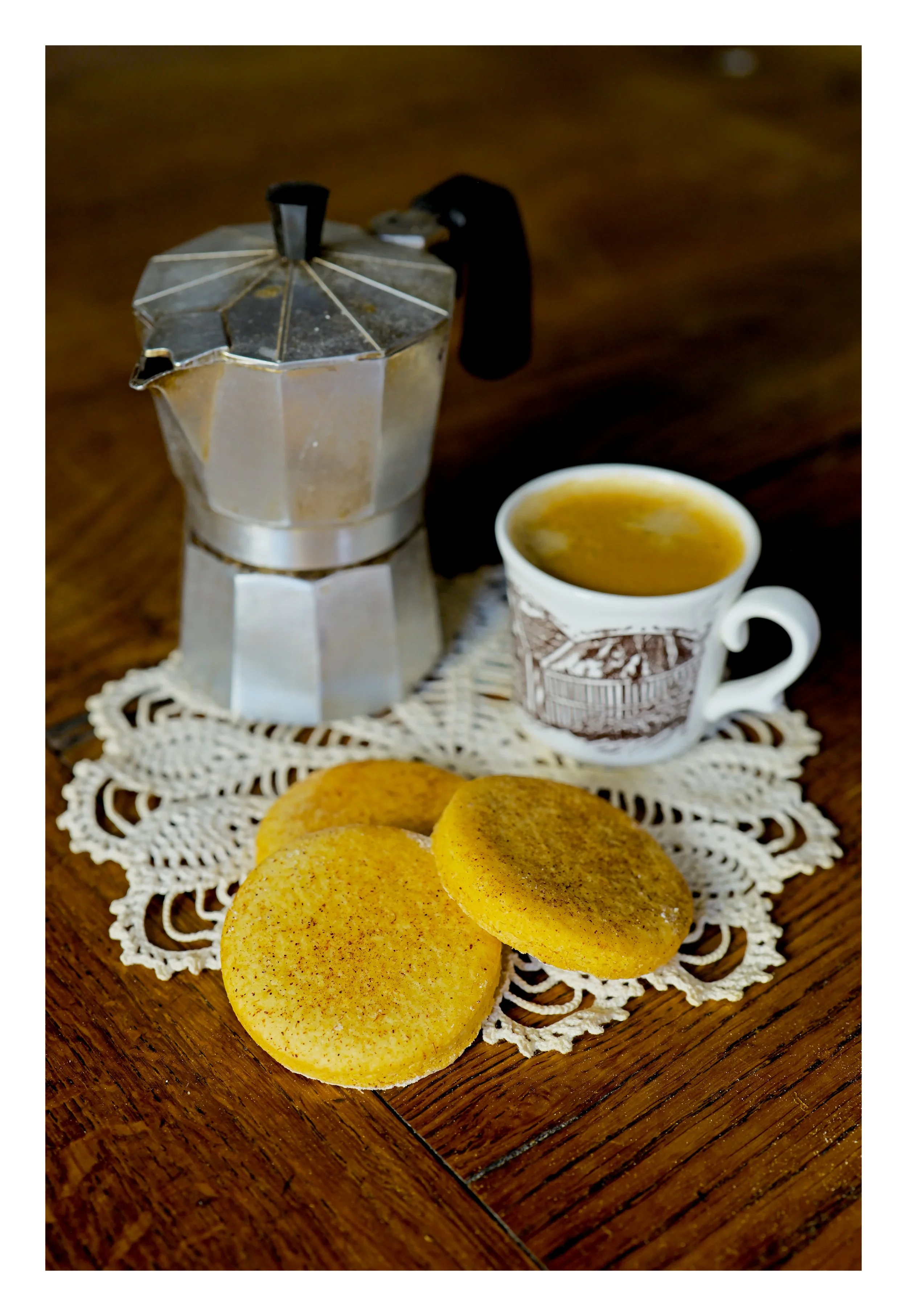 Un moka en aluminium, une tasse de café, des cookies, et une nappe en dentelle sur une table en bois.