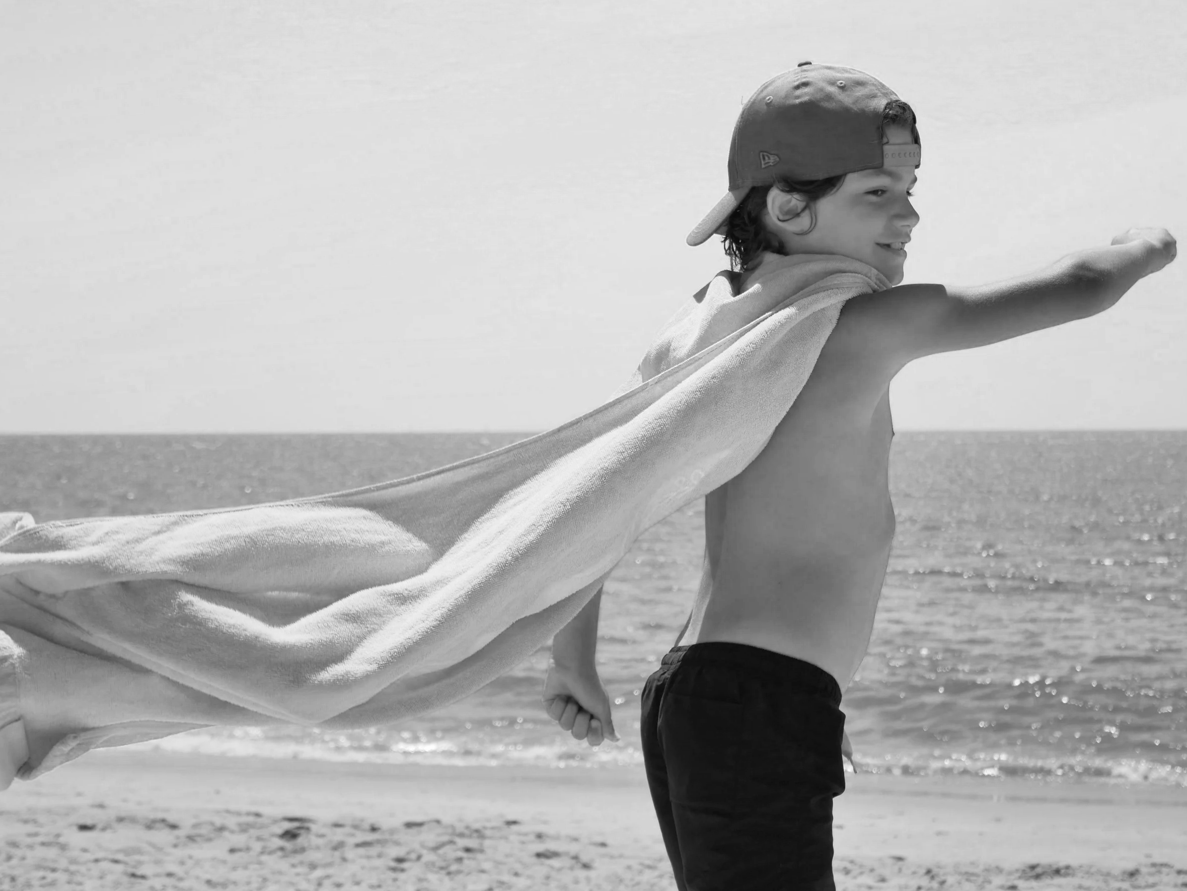 Un garçon avec une casquette portée à l'envers marche sur la plage en tenant une serviette ou un vêtement qui flotte dans le vent.