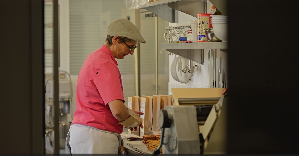 Une femme porte un uniforme rose et une toque, elle emballe des cookies dans une cuisine de pâtisserie.