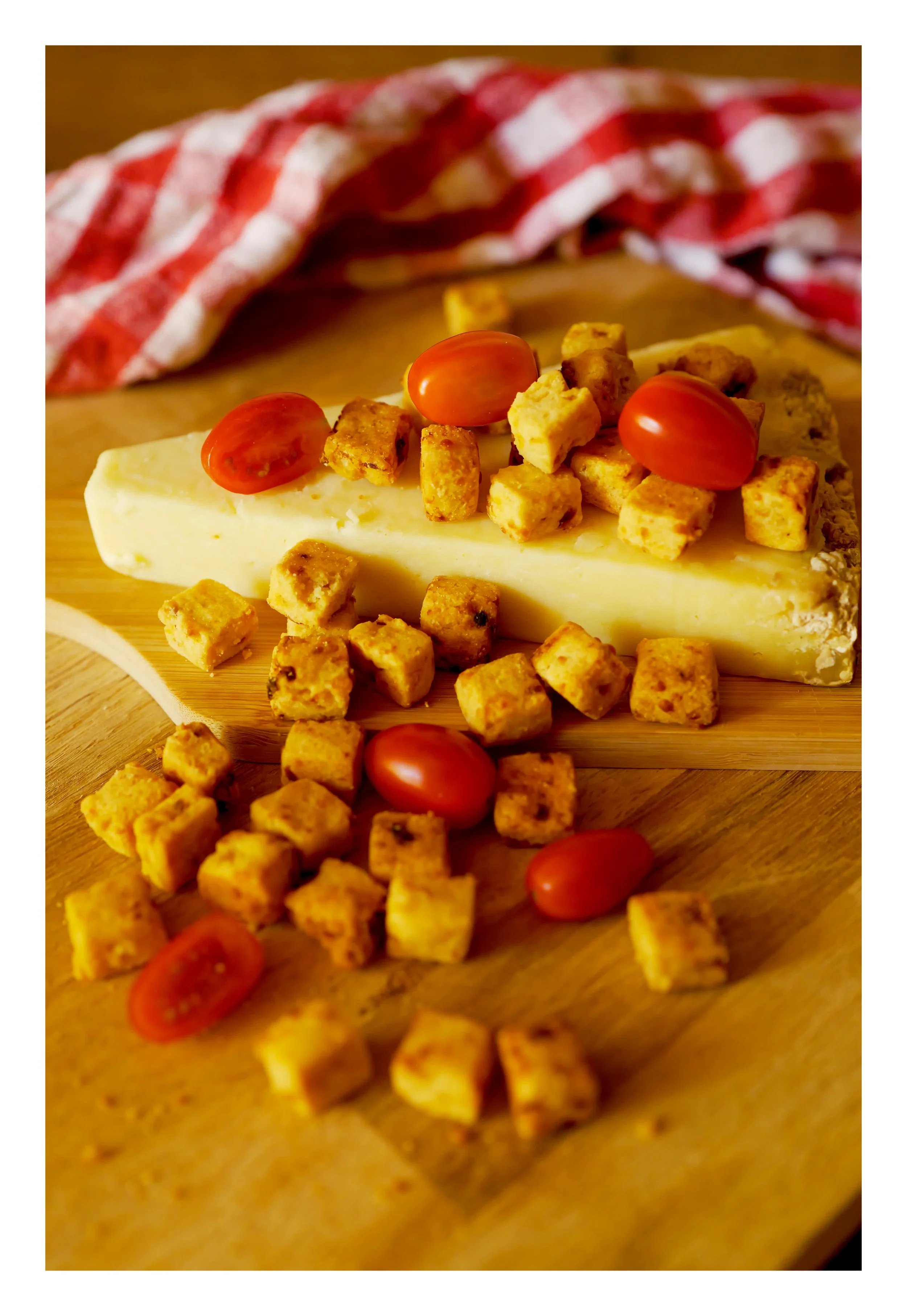 Un morceau de fromage blanc avec des tomates cerises rouges, des croûtons dorés, posé sur une planche en bois, avec un torchon à carreaux rouge et blanc en arrière-plan.