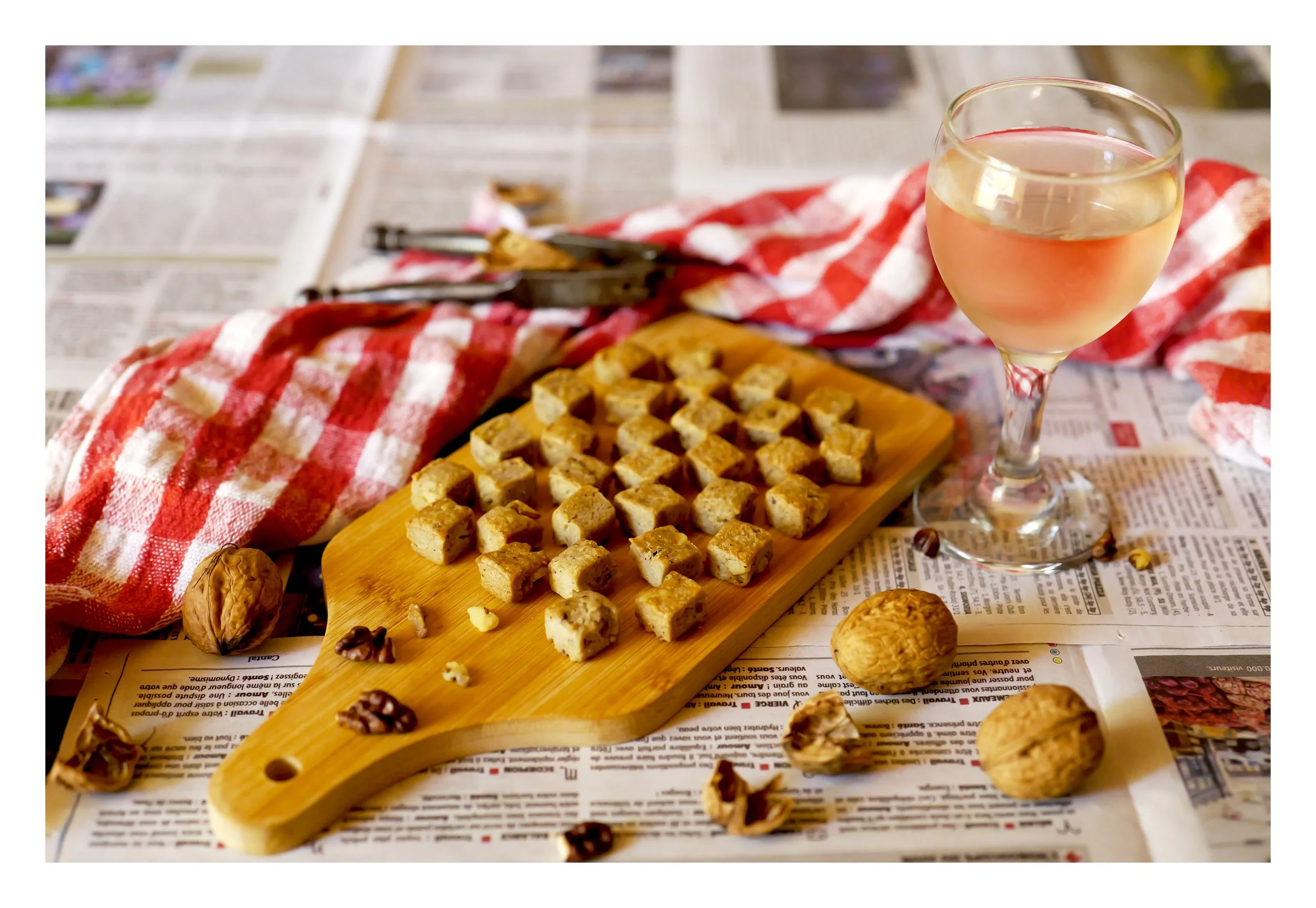 Une coupe de vin blanc à côté d'un plateau de petits biscuits, le tout posé sur une table recouverte de journaux et d'une serviette à carreaux rouges et blancs.