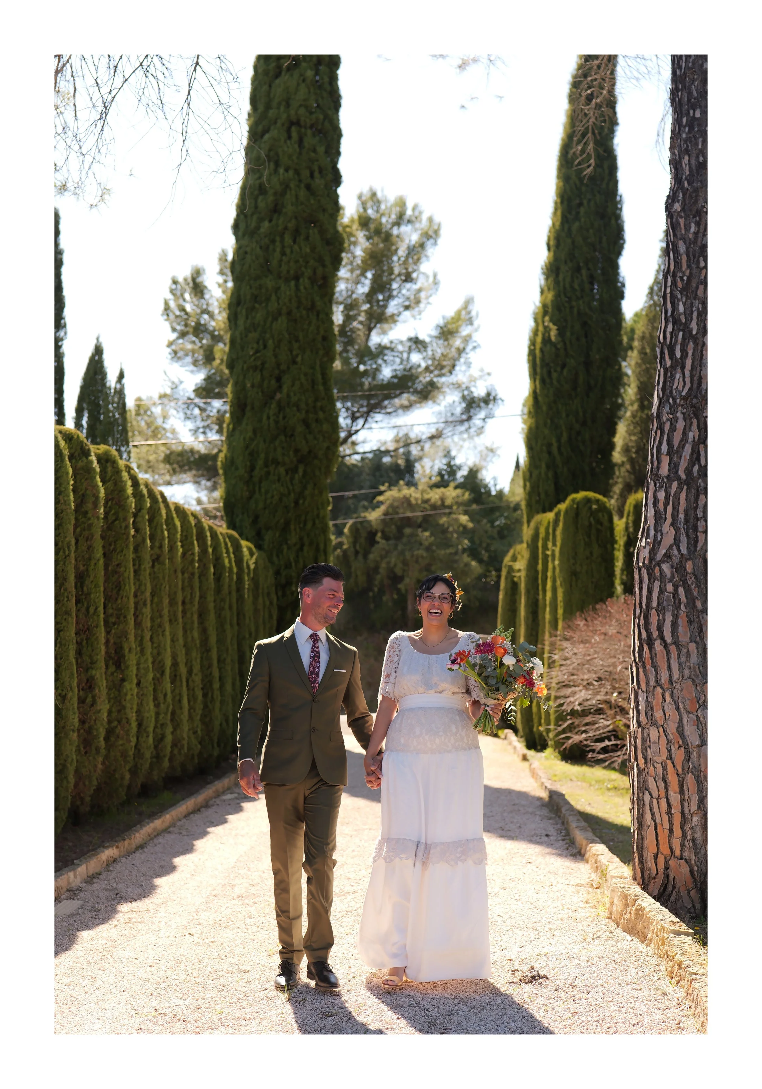 Un couple marchant main dans la main dans un jardin, la femme porte une robe de mariée blanche avec un bouquet de fleurs, l'homme porte un costume vert. Ils semblent heureux et souriants.