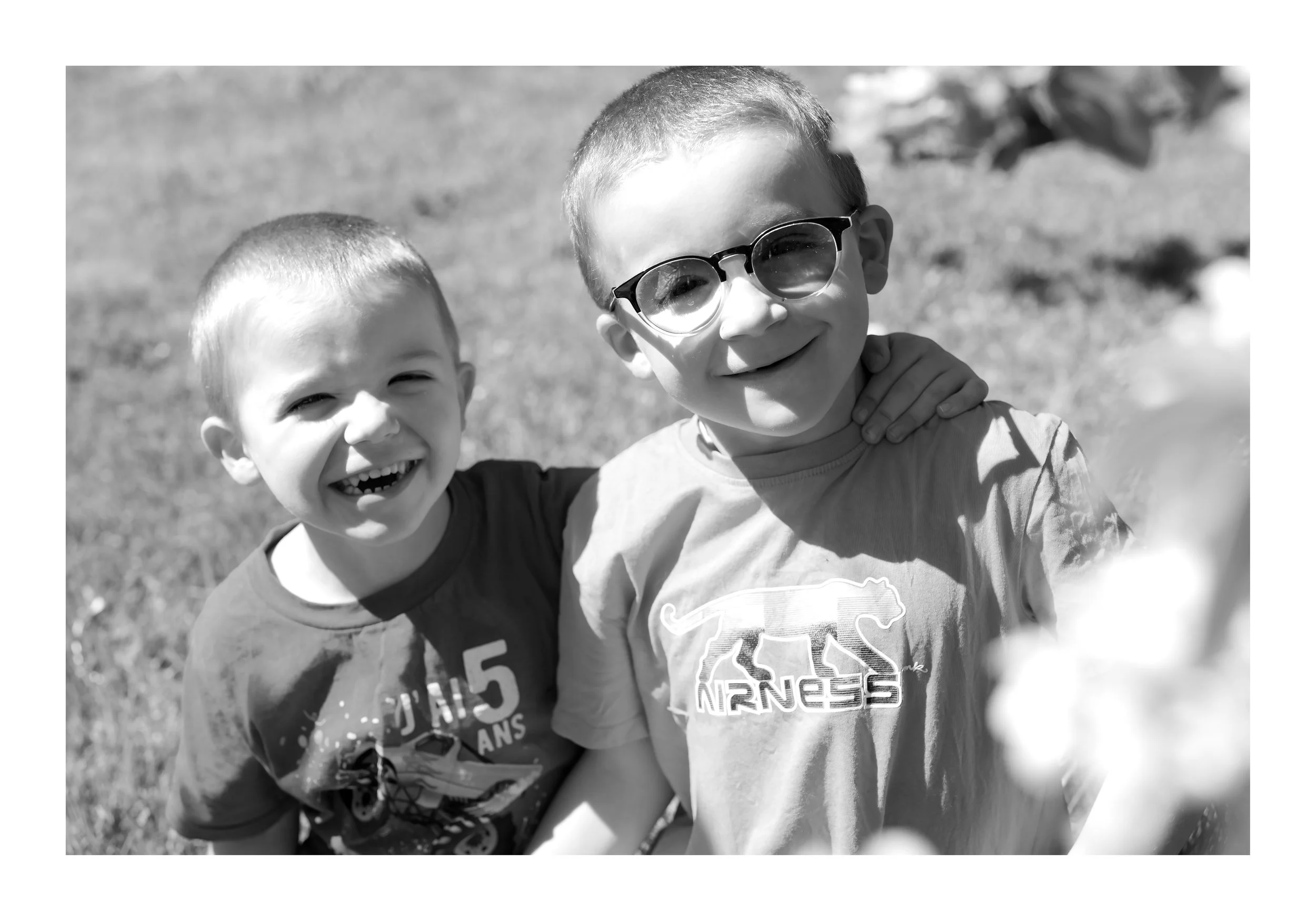 Deux enfants souriants en extérieur, l’un porte des lunettes et l’autre un t-shirt avec un dessin de route.
