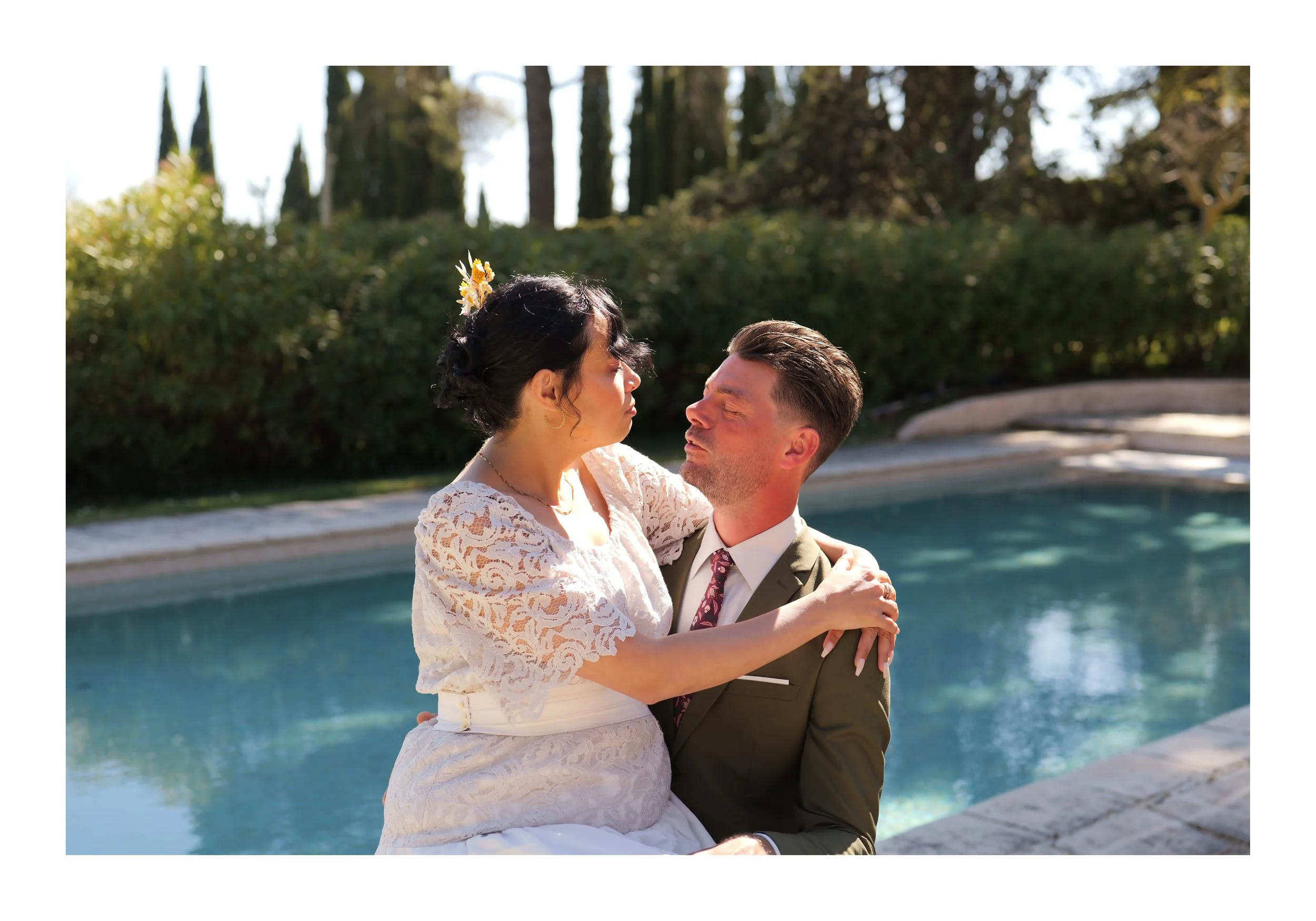 Un couple en tenue de mariage, la femme portant une robe en dentelle blanche et un petit bouquet dans les cheveux, le homme en costume, se regarde tendrement près d'une piscine extérieure ensoleillée avec un fond de végétation et d'arbres.