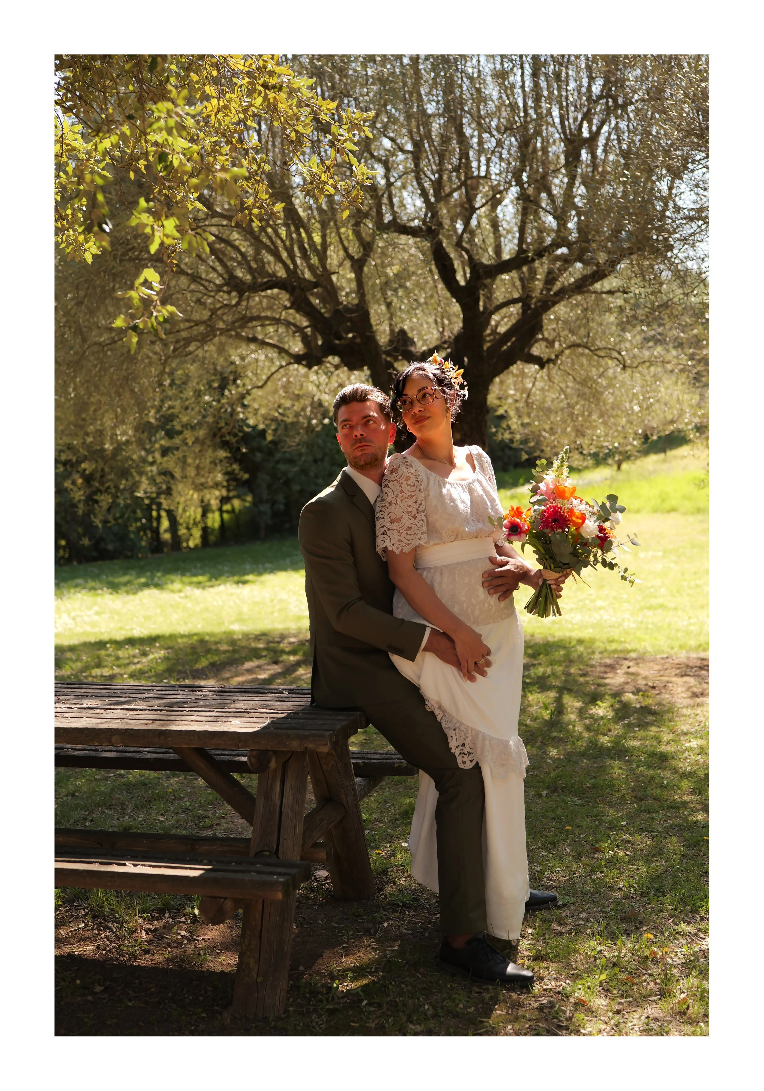 Un couple en tenue de mariage assis sur un banc en bois dans un parc, entouré d'arbres, avec la femme tenant un bouquet de fleurs colorées.