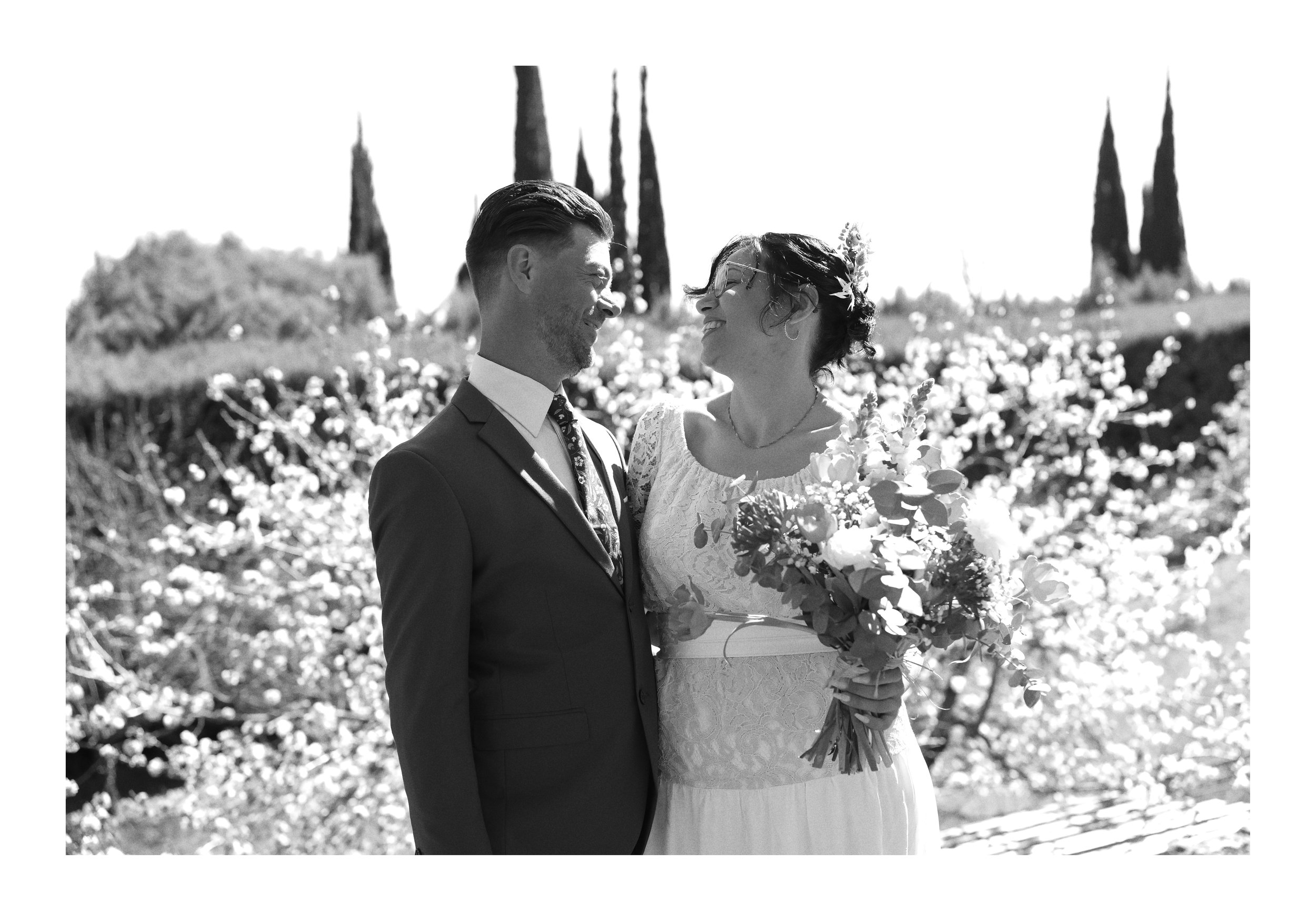 Un couple de mariés souriants lors de leur mariage en plein air, avec des arbres en arrière-plan et des fleurs dans la main de la mariée.