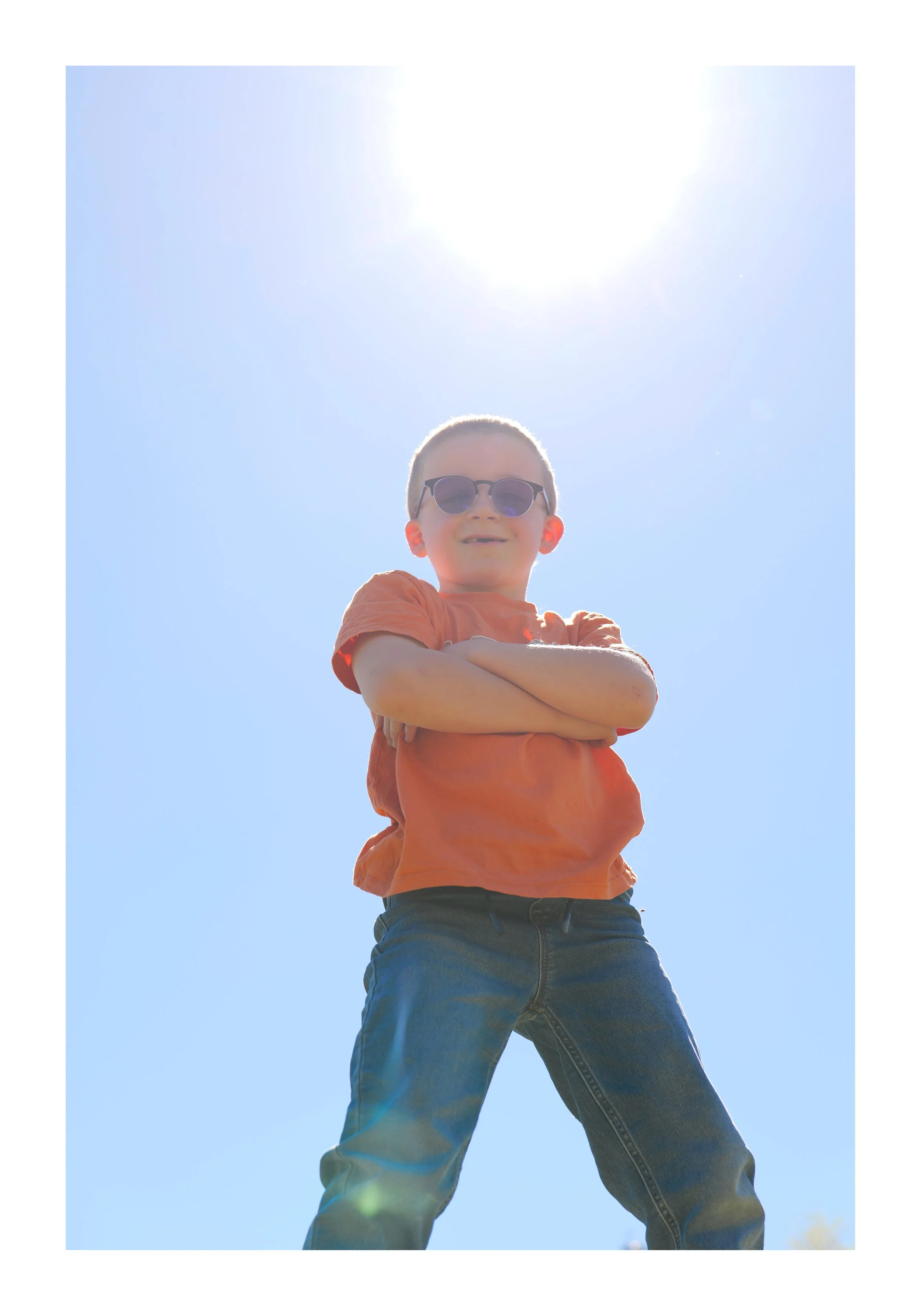 Un garçon portant des lunettes de soleil, un t-shirt orange, et un jeans, croisé les bras, se tenant debout sous un ciel bleu avec le soleil brillant au-dessus.