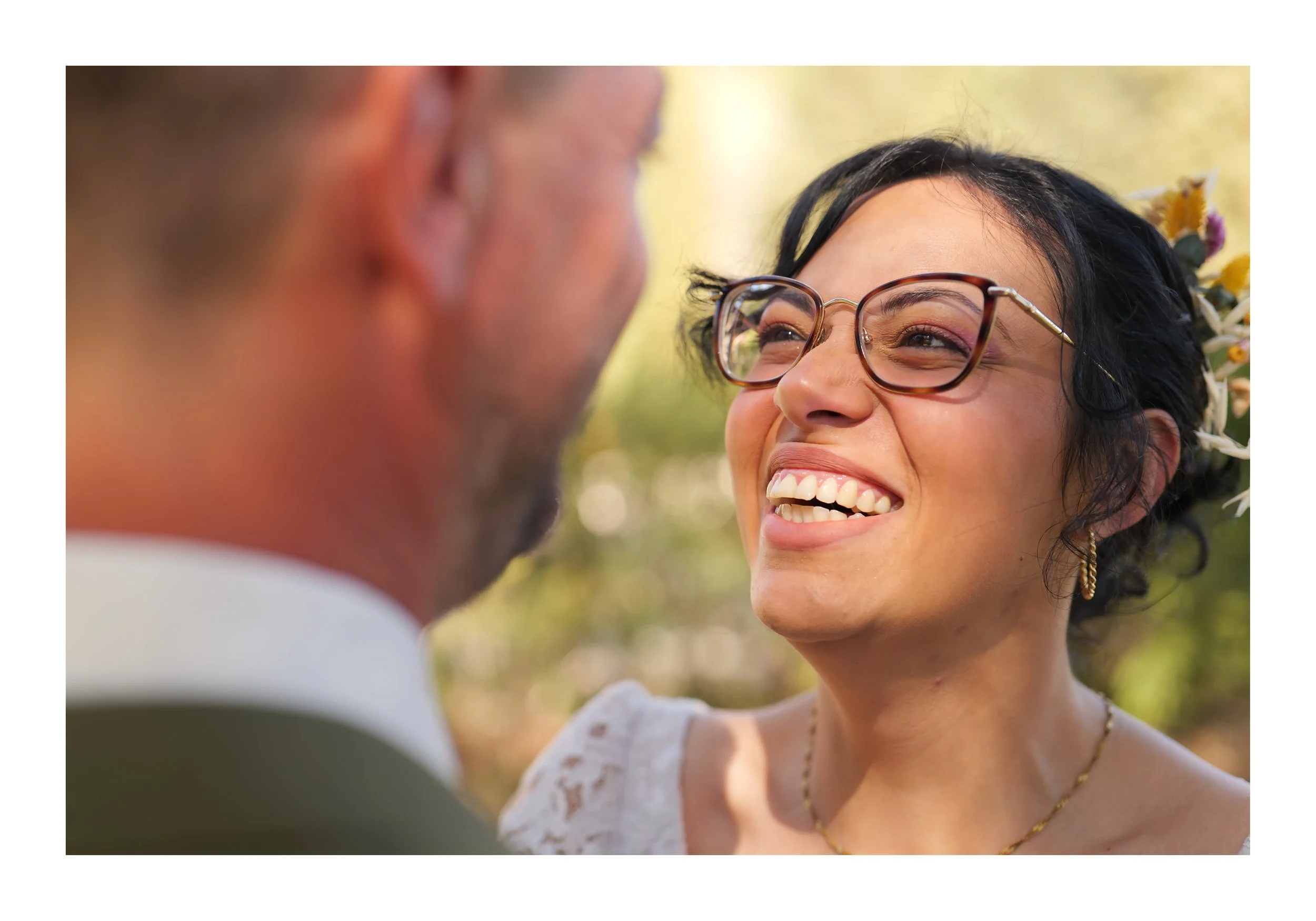 Une femme souriante avec des lunettes et des cheveux noirs, portant des bijoux dorés et un vêtement blanc, regarde un homme qui est flou en premier plan. Le fond est un environnement extérieur lumineux et fleuri.