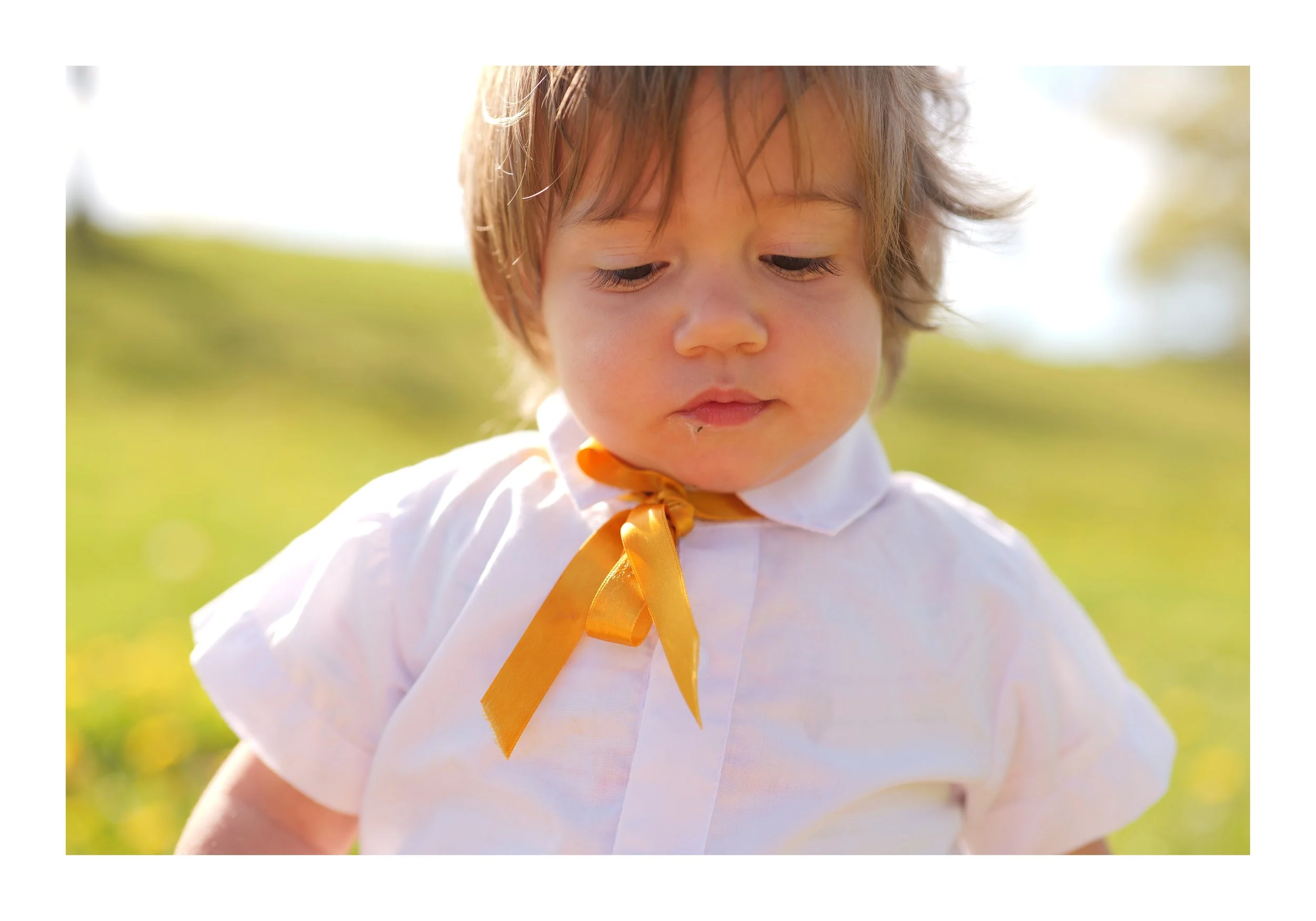 Un jeune garçon avec des cheveux bruns, portant une chemise blanche et un ruban orange autour du cou, regarde vers le bas à l'extérieur avec un fond végétal flou.