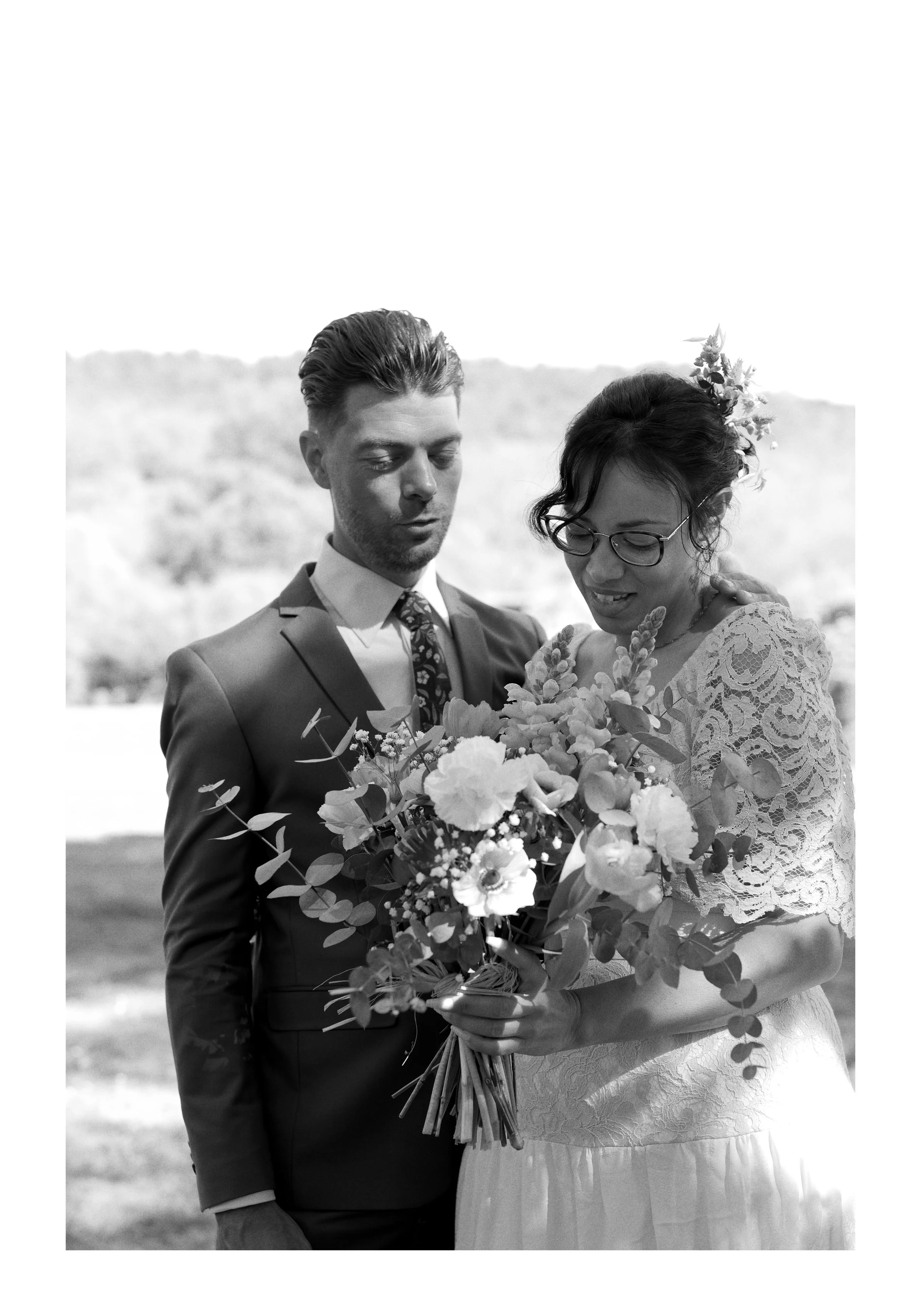 Un couple de mariés lors d'un mariage en plein air, la femme porte une robe en dentelle et des lunettes, tenant un bouquet de fleurs, et l'homme porte un costume.