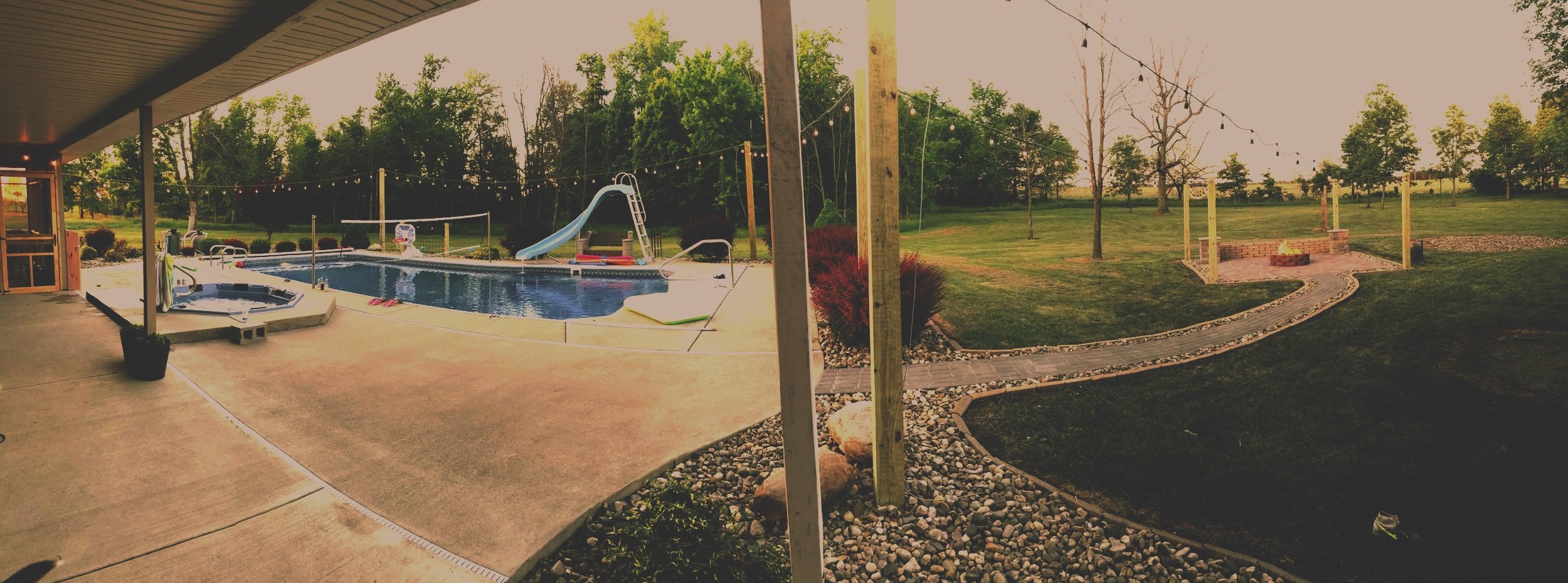 A backyard with a concrete patio, a swimming pool with a slide and a hot tub, surrounded by a grassy lawn with trees and a fire pit area under string lights.