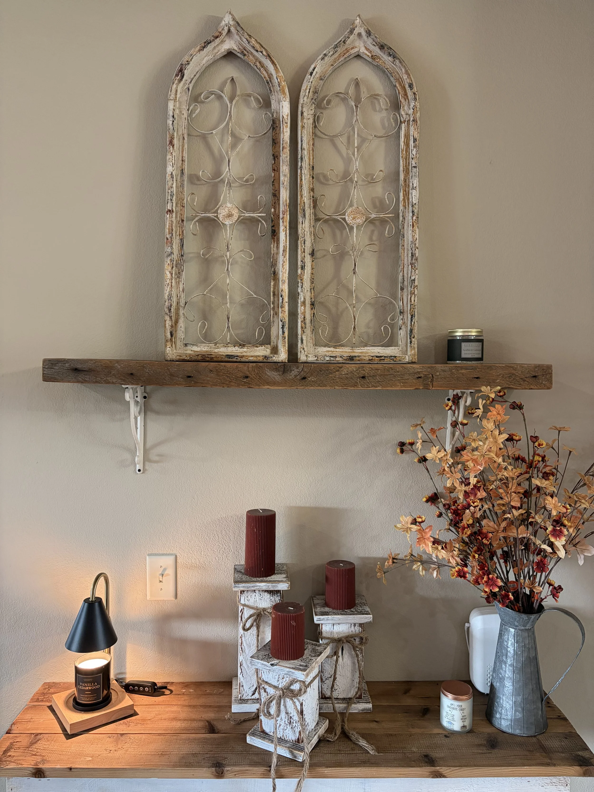 Decorative wall display with two rustic, distressed arched frames with intricate metal scrollwork, a wooden shelf below, three red candles on wooden crates, a black modern desk lamp, a large metal vase with artificial orange autumn leaves, and a small container holding a picture and some text.