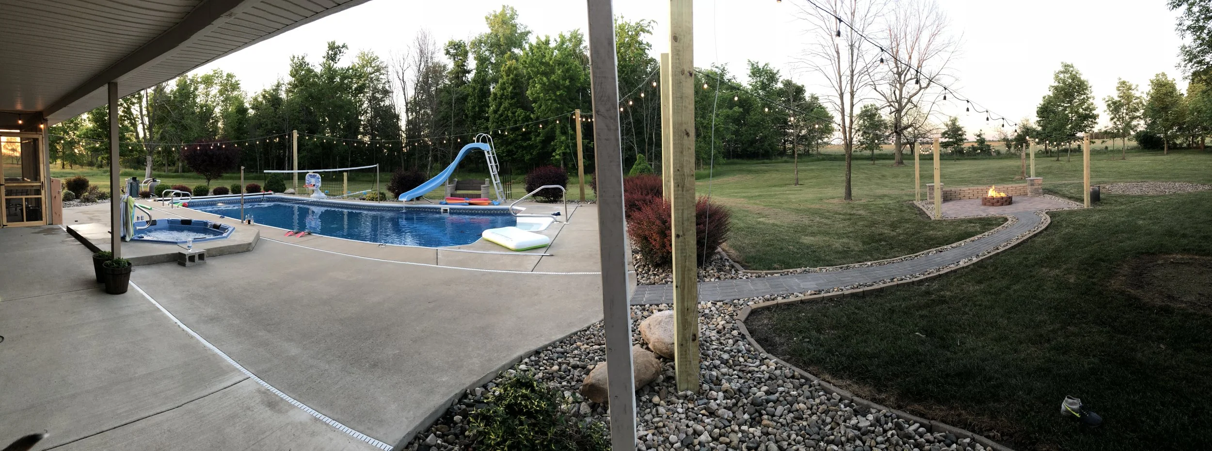 A backyard with a swimming pool, lounge float, and a slide surrounded by grass and trees, with a fire pit and string lights.