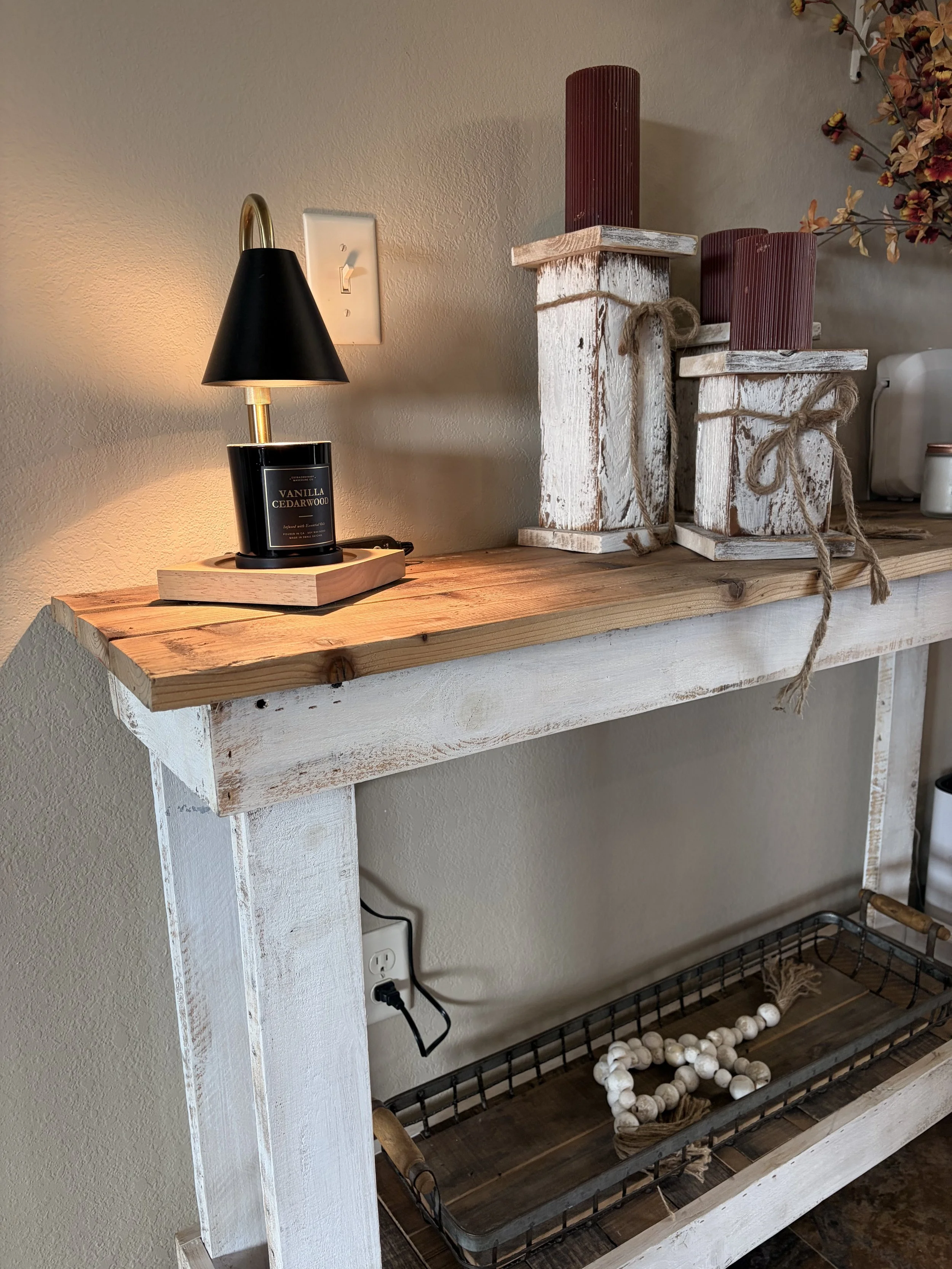 A rustic wooden shelf decorated with a black table lamp with a gold arm and a vanilla cedarwood scented candle, alongside decorative items including large white beads, wrapped twine, and red wooden vases. The shelf is placed against a beige wall.