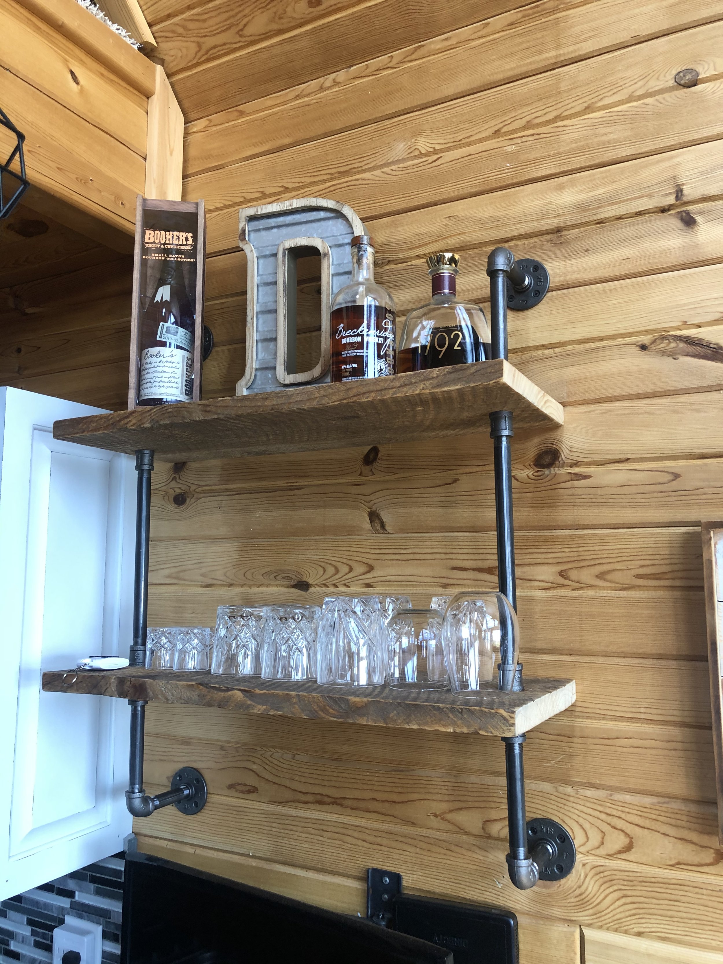 Wooden wall with three wooden shelves supported by black metal pipes, holding glassware, whiskey bottles, a decorative letter 'D', and a book titled 'Booker's' in a cozy kitchen setting.
