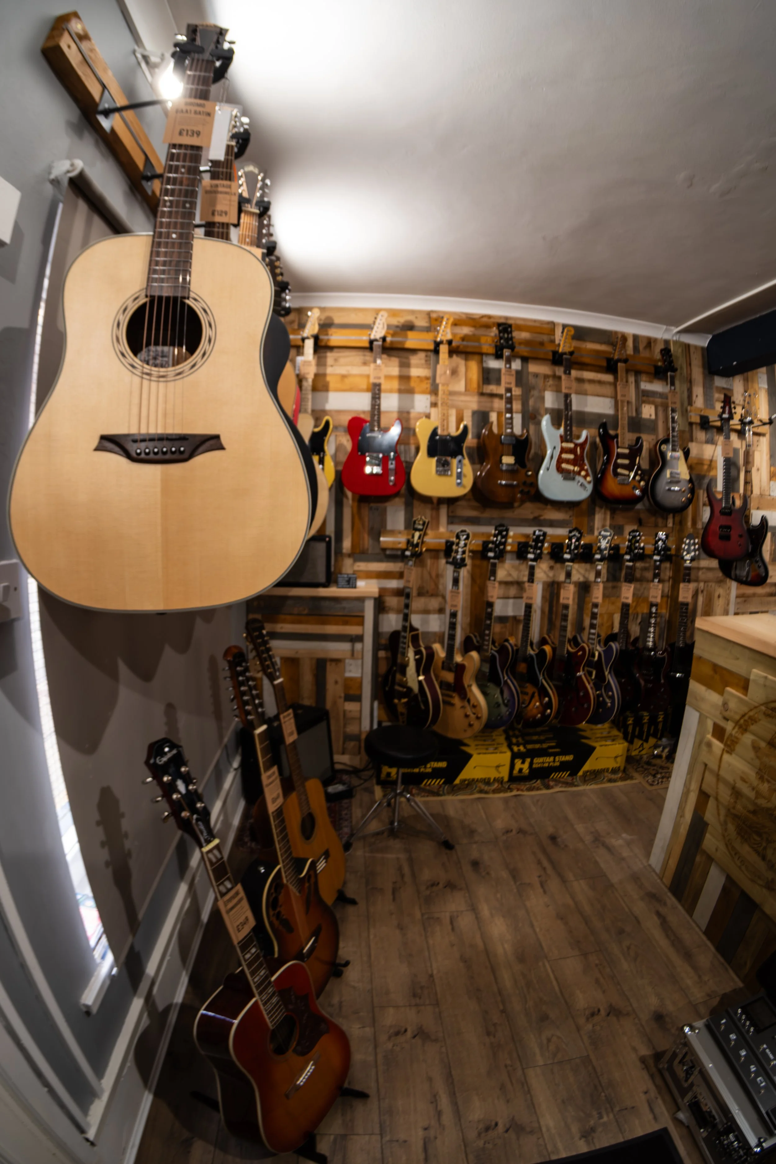 A music store displays various acoustic and electric guitars on walls and stands, with some guitars hanging and others on guitar stands on the floor, in a cozy wooden store.