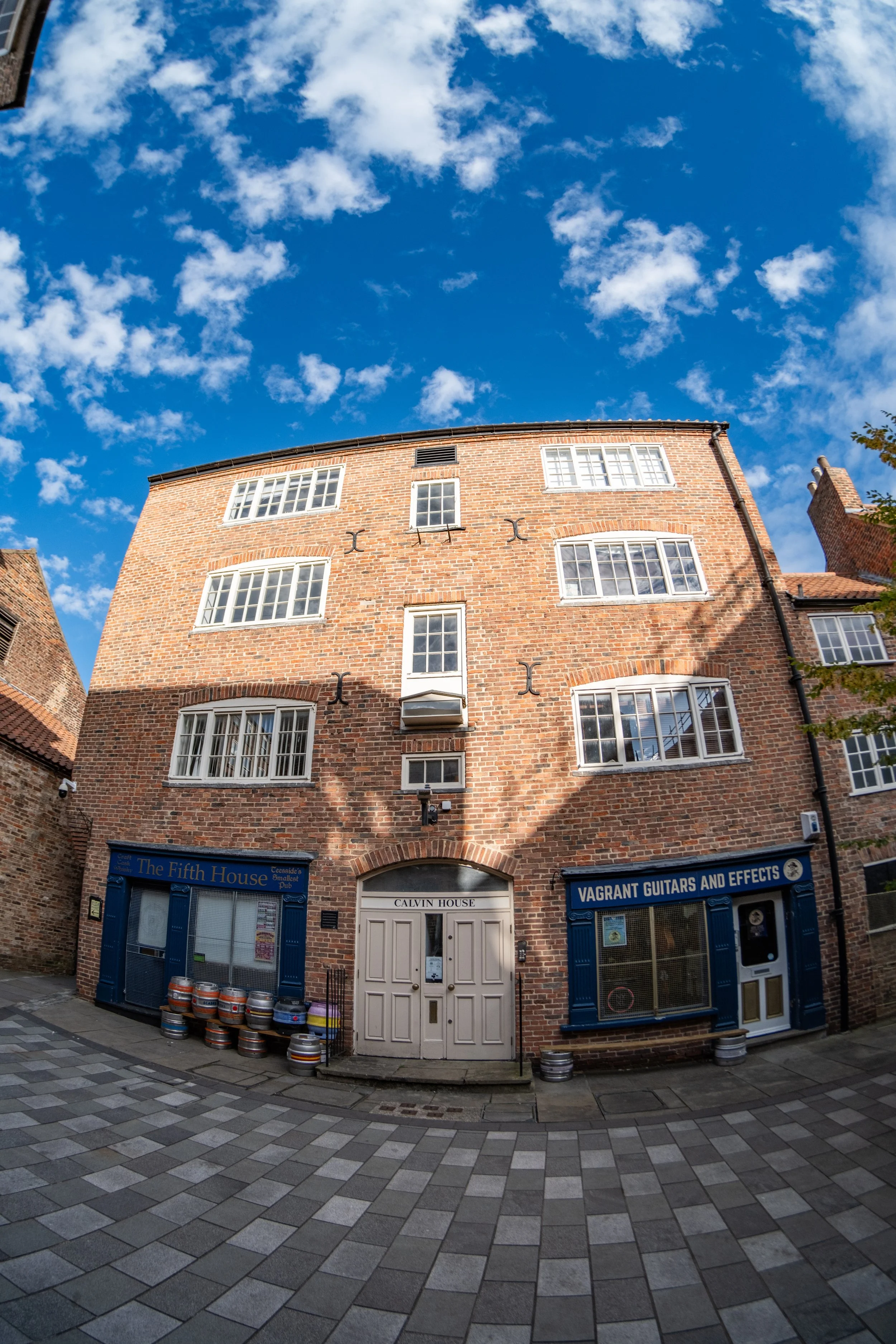 The image shows a multi-story brick building with blue storefronts at street level. The storefronts have signs that read "The Fifth House" and "Vagrant Guitars and Effects." The building has several white-framed windows on each floor. The sky above is blue with scattered white clouds, and the street in front is paved with gray and beige tiles.