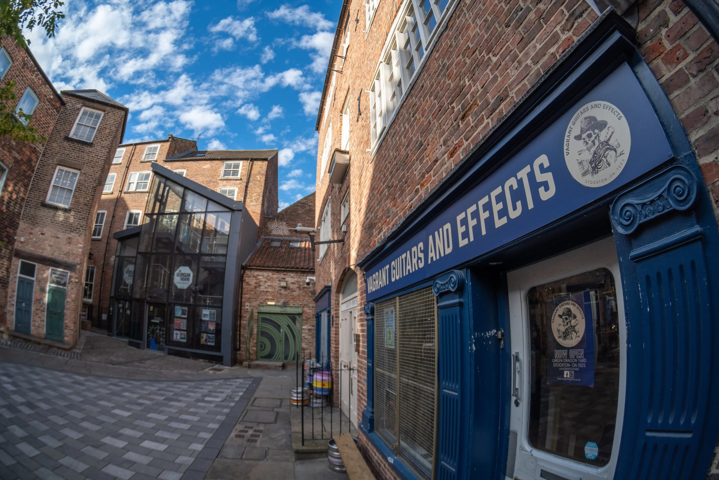 Street view of brick buildings with a guitar shop called "Vagrant Guitars and Effects" on the right. The shop has a blue façade with a sign and a circular logo featuring a skeleton wearing a hat. The street has a patterned sidewalk, and there are barrels and a green arched door nearby. The sky is partly cloudy with blue sky showing.