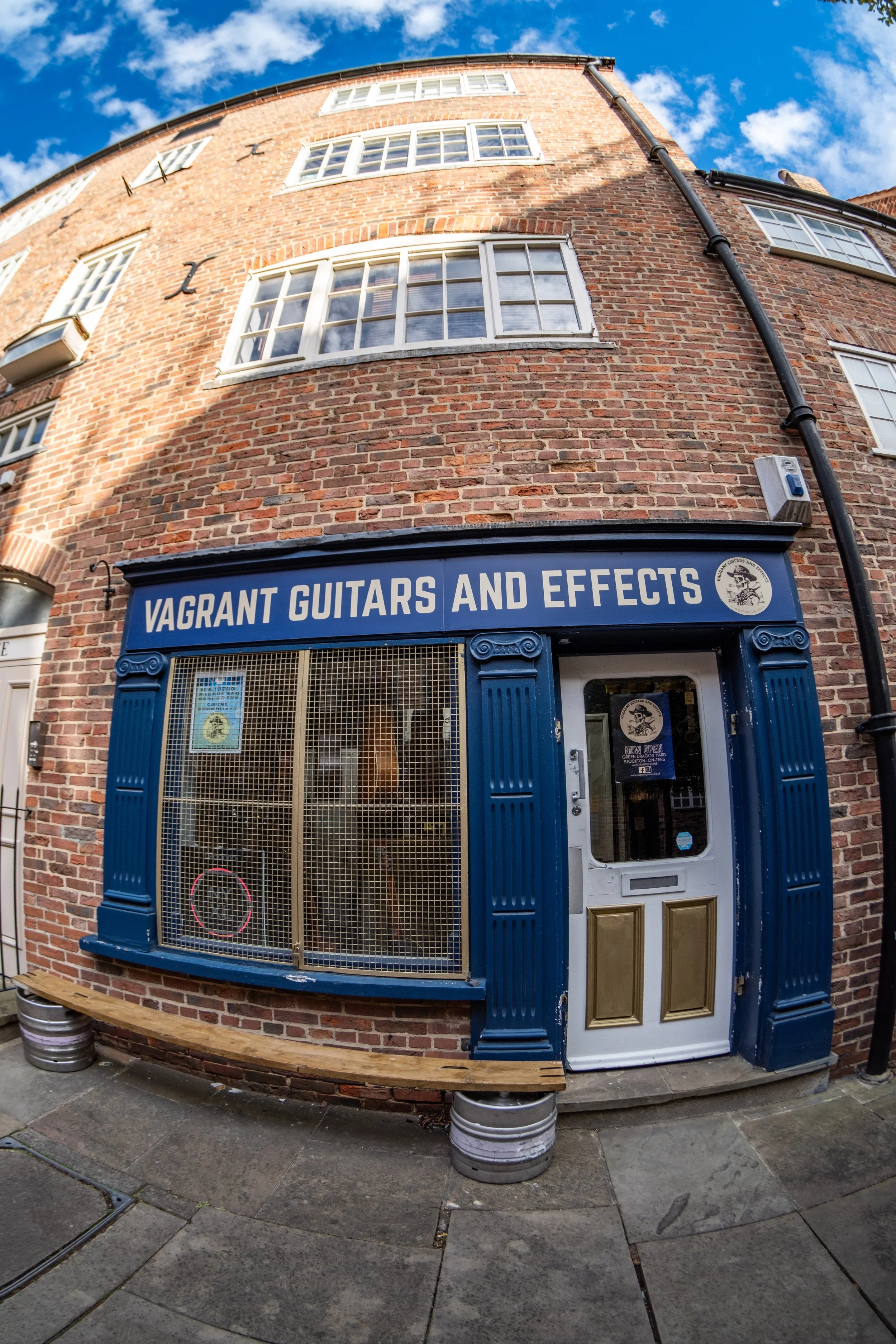 A storefront with a blue sign reading 'Vagrant Guitars and Effects' on a brick building, with a large window display and a white door, under a bright blue sky with scattered clouds.