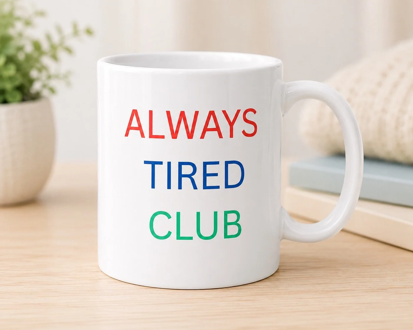 White mug with the words 'ALWAYS TIRED CLUB' printed in red, blue, and green letters, placed on a wooden surface with a plant and books in the background.