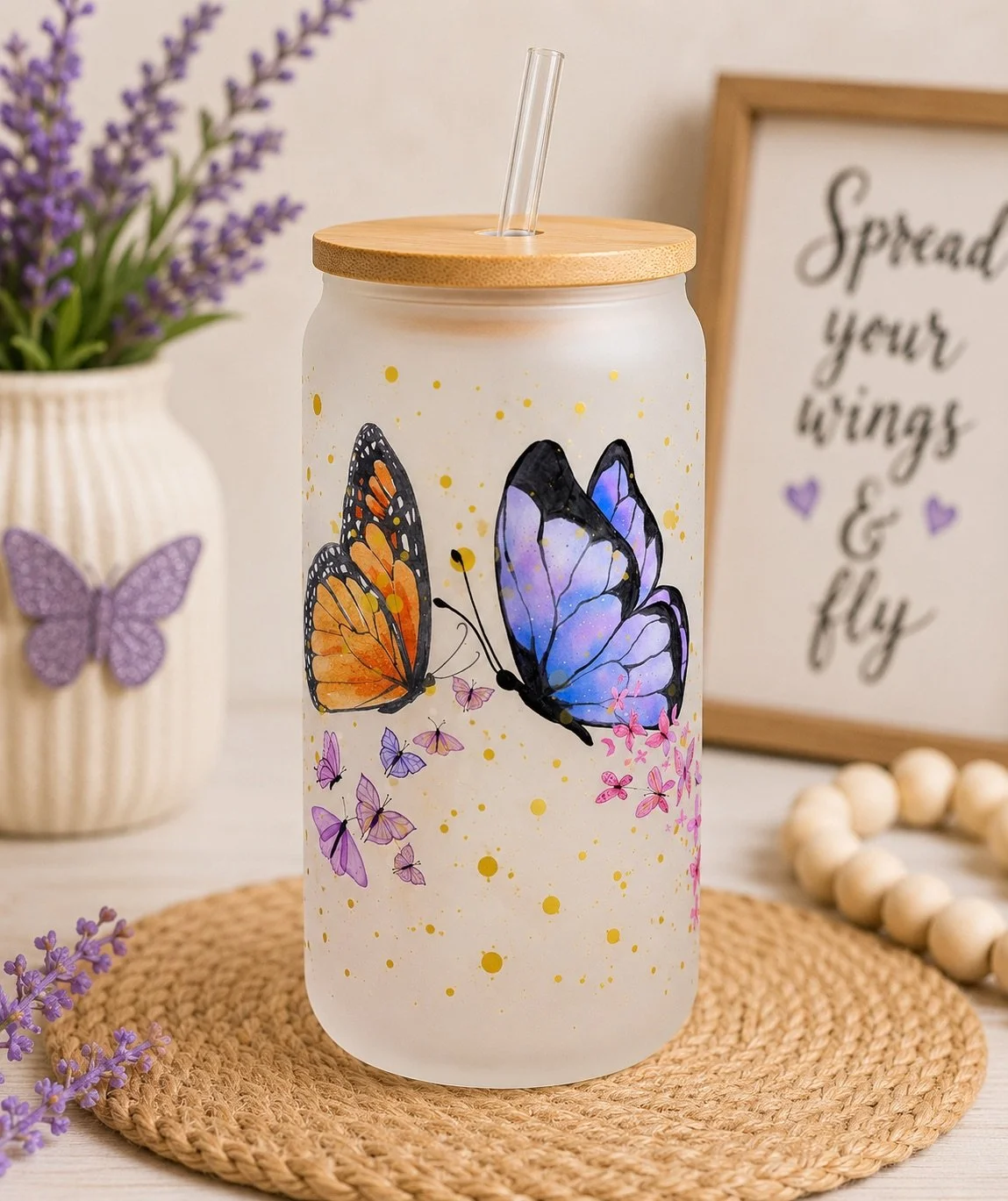 Decorative frosted glass tumbler with painted orange and purple butterflies, gold splatters, a bamboo lid, and a straw, placed on a woven placemat with lavender flowers and a butterfly decoration nearby, and a framed quote in the background that says "Spread your wings & fly."