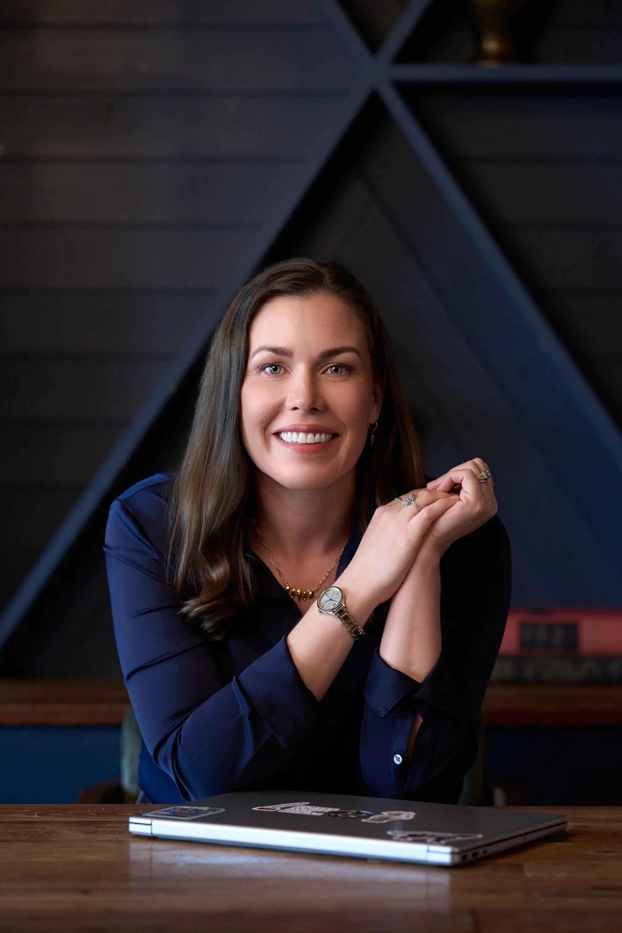 A woman with long brown hair and blue eyes smiling, wearing a navy blazer, gold jewelry, and a watch, sitting at a wooden table with a closed laptop and some stickers on it, in a dimly lit room with black paneled walls.