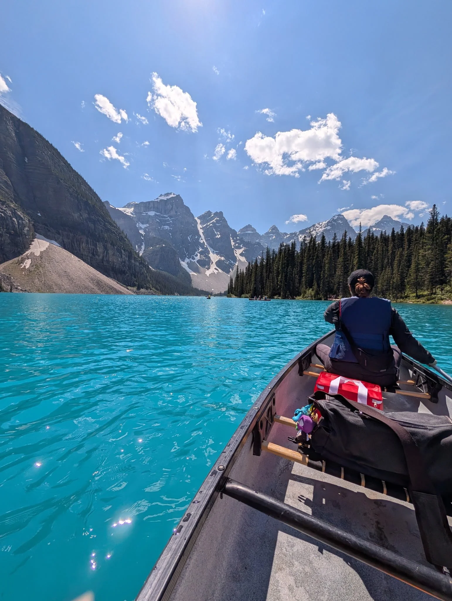 A person sitting in a canoe on a turquoise lake surrounded by mountains with snow patches and a forest of pine trees, under a partly cloudy blue sky.
