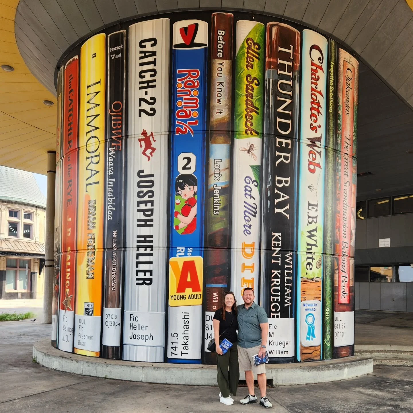 A large outdoor art installation resembling a row of oversized colorful books with titles and authors' names, with a smiling couple standing in front of it.