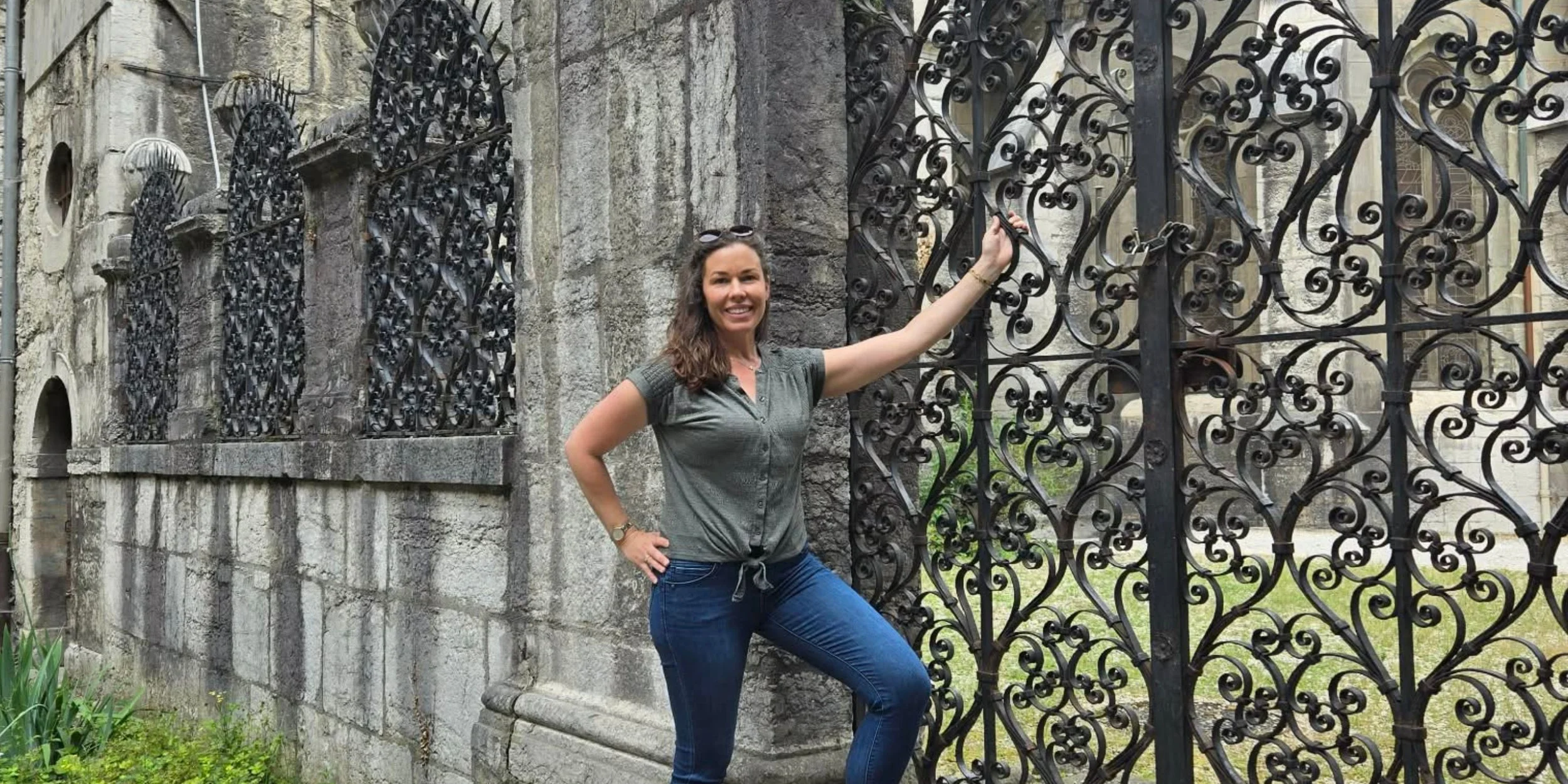 A young woman in a green shirt and jeans is standing in front of an old ornate church gate with one hand on her hip and the other on the gate.