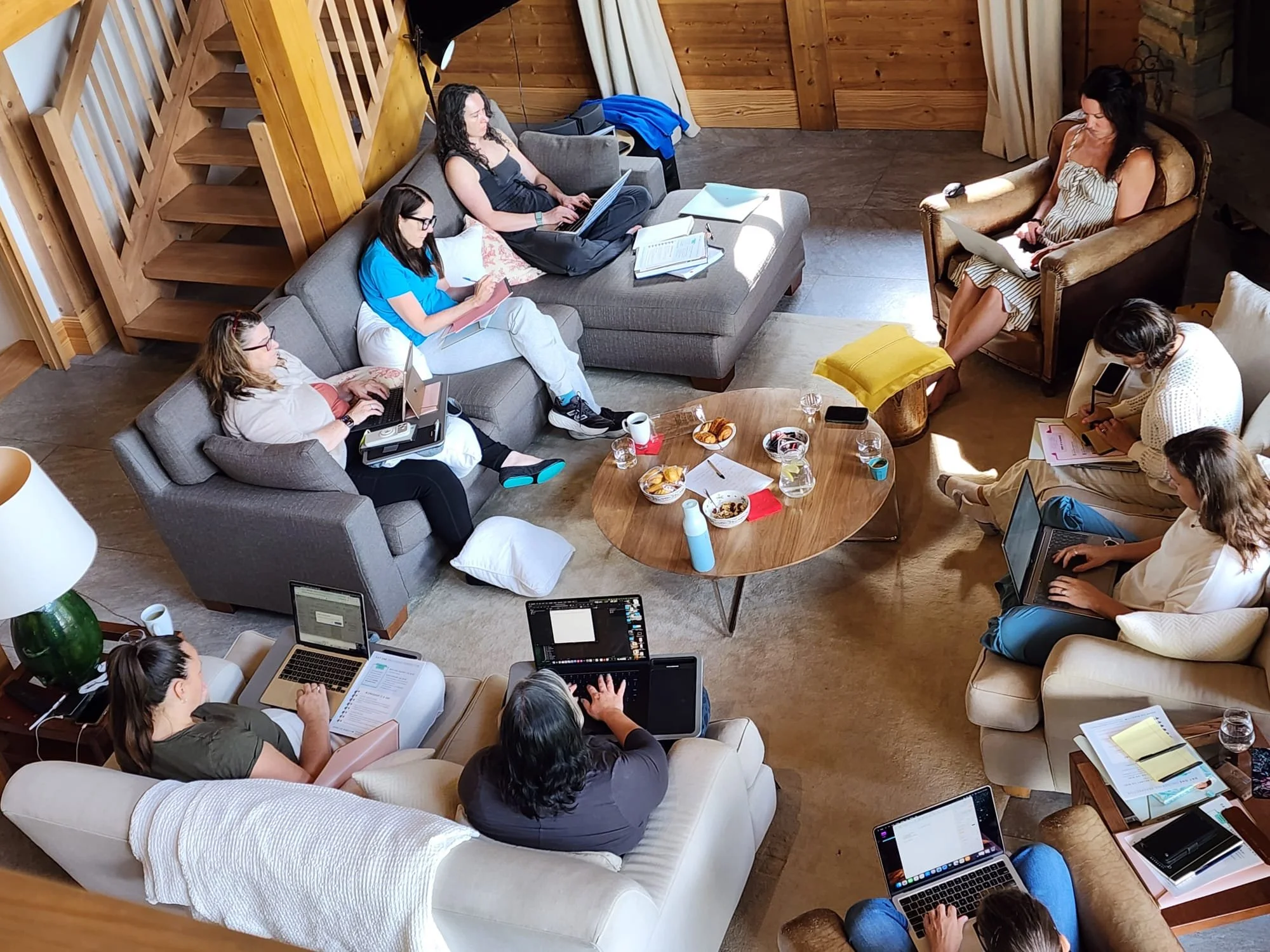 A group of people sitting in a cozy living room with wooden walls, engaged in a meeting or collaborative work, with laptops, notebooks, and snacks on a coffee table.