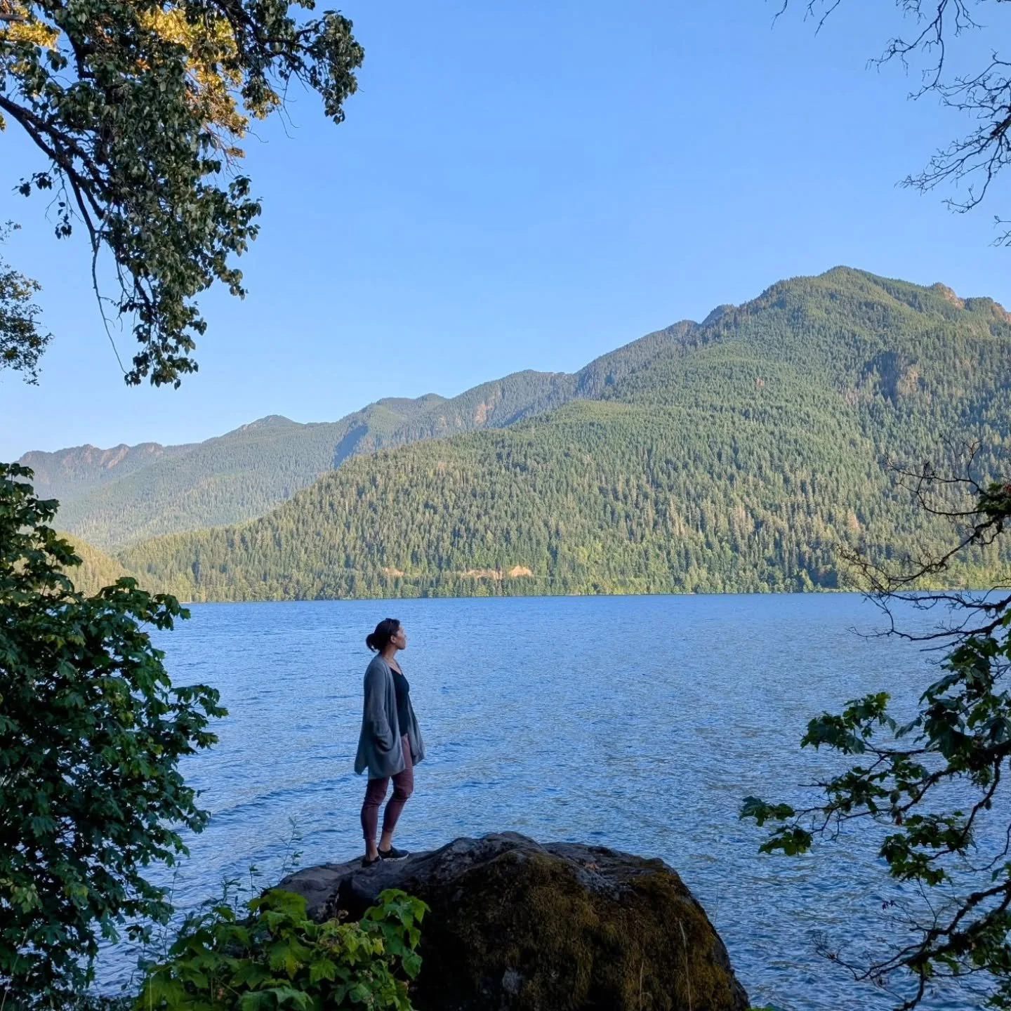 A woman stands on a large rock by a lake, surrounded by trees, with mountains in the background under a clear blue sky.
